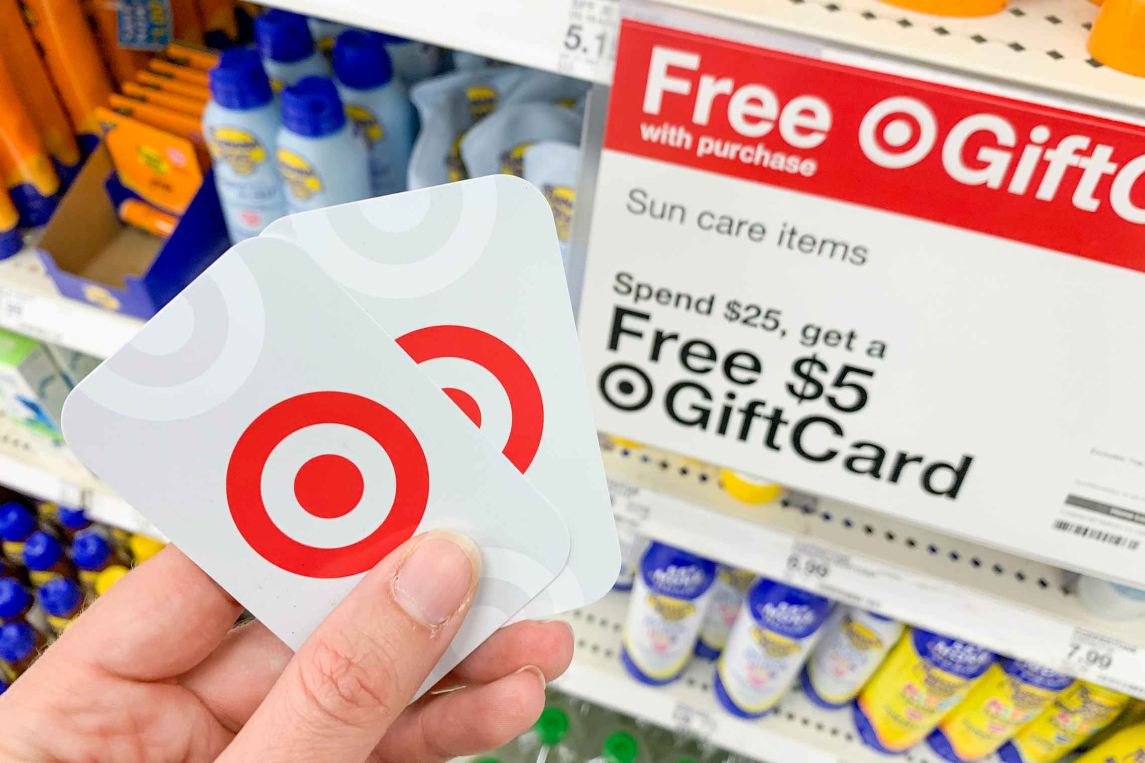 A person's hand holding two Target gift cards in front of a sign advertising getting a free Target gift card when you spend $25 on sun care items.