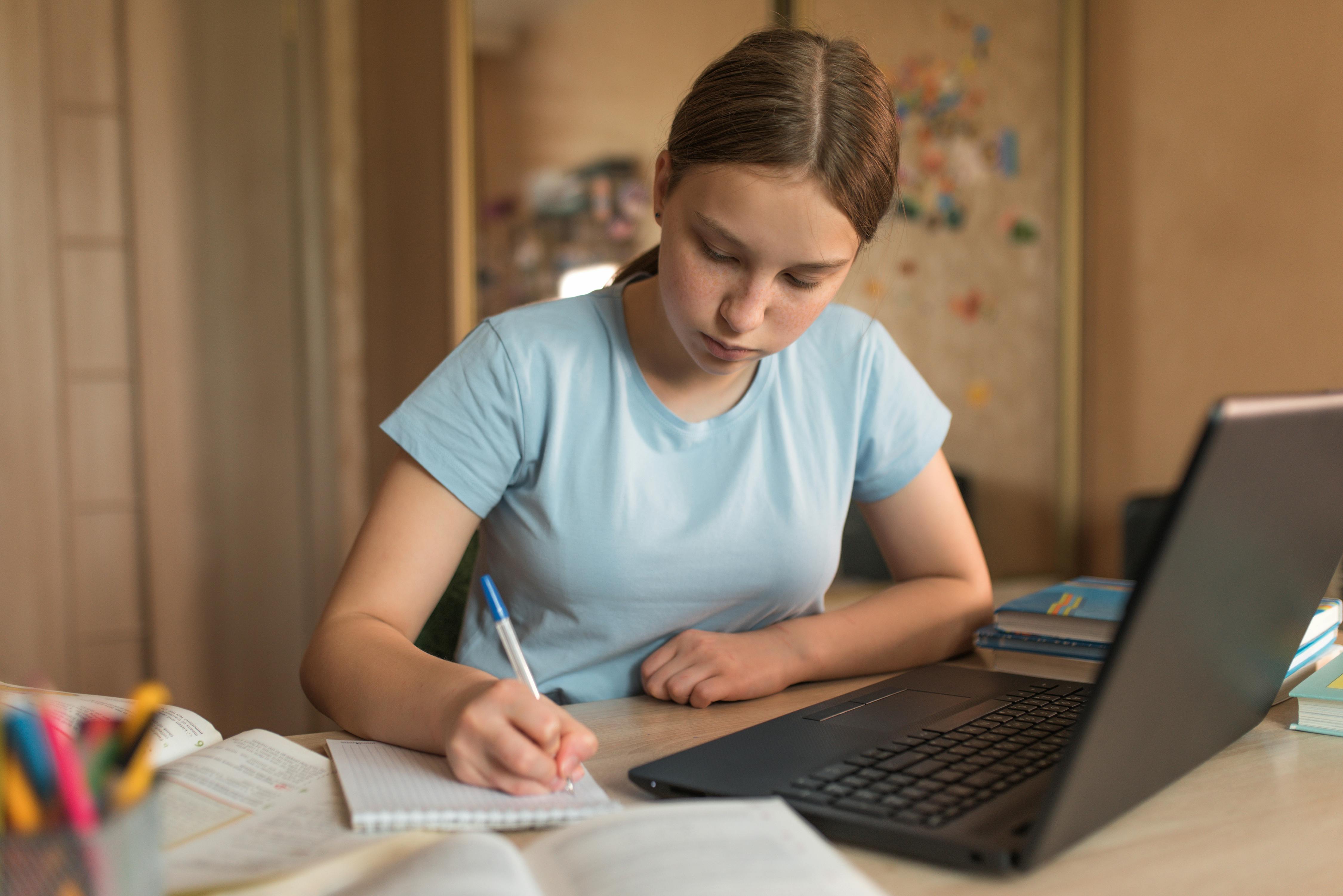 A teen girl sitting at a desk with some books and a laptop, writing in a notebook.