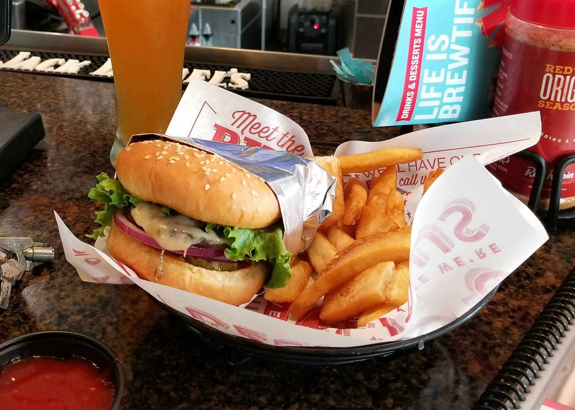 A Red Robin burger in a basket with fries sitting on a table next to a dip cup of ketchup and a beer glass.