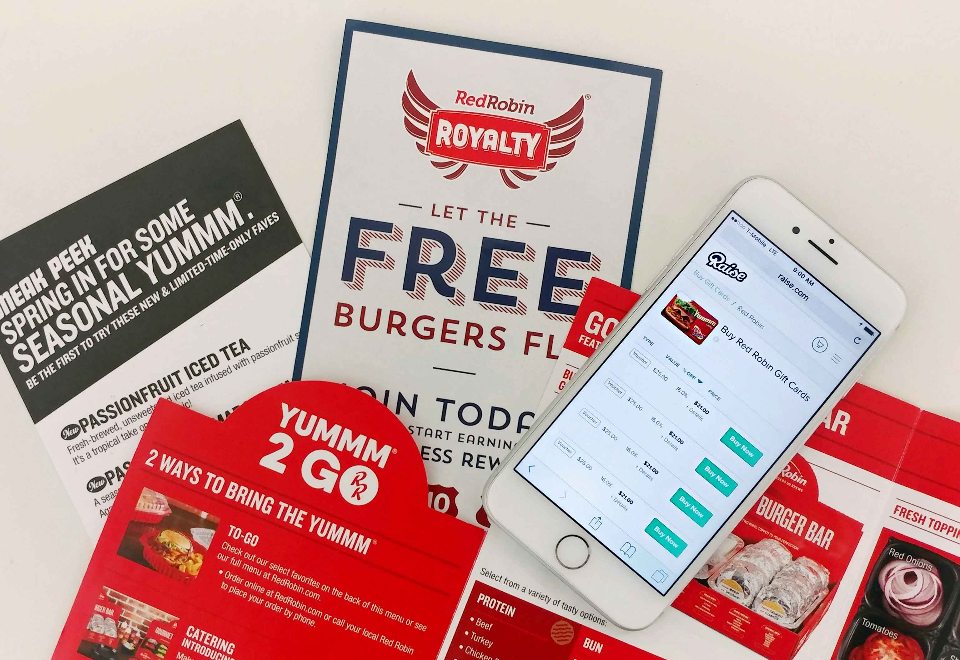 A phone displaying Raise.com's Red Robin gift card page laying on a table with Red Robin menus and leaflets for the Royalty Rewards program.