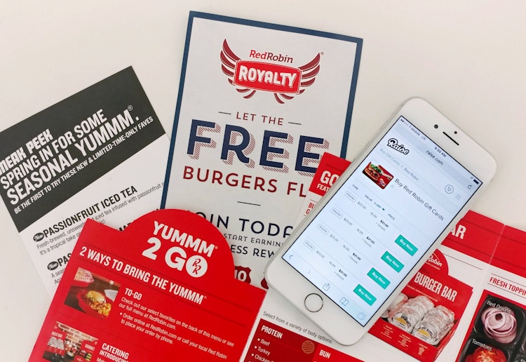 A phone displaying Raise.com's Red Robin gift card page laying on a table with Red Robin menus and leaflets for the Royalty Rewards program.