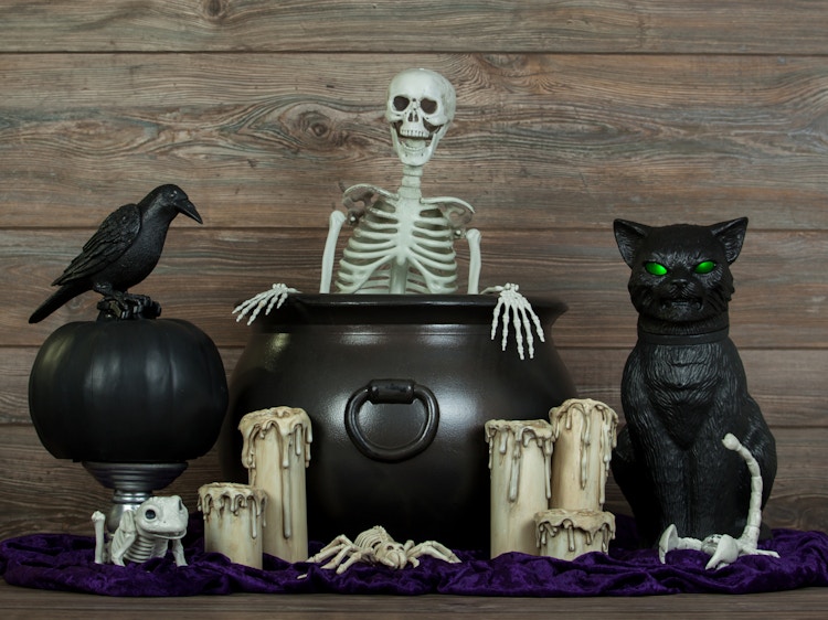 Halloween decorations grouped together on a table including a cauldron, a skeleton, a crow, a black cat, some candles, and more.