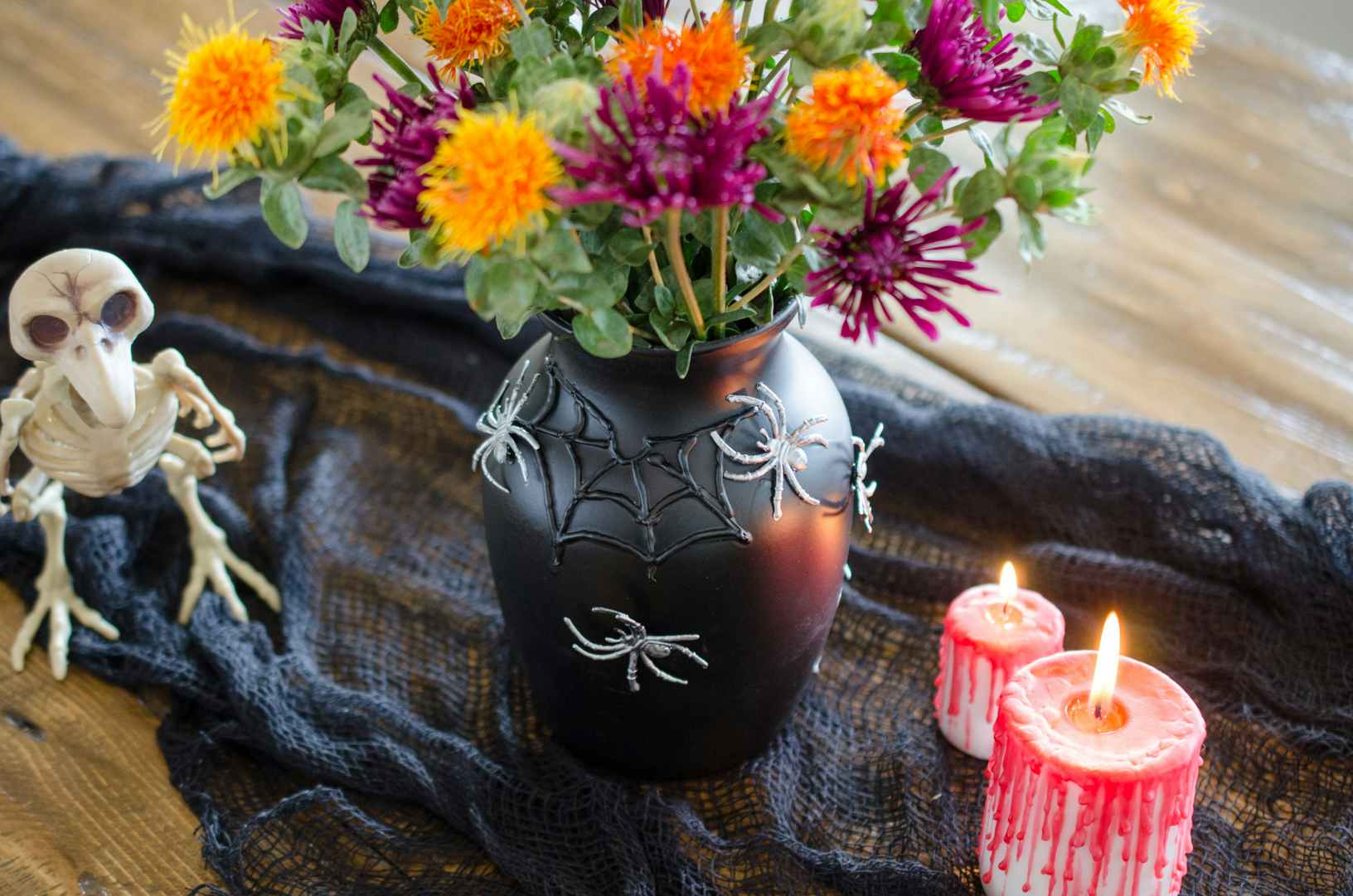 A black vase covered in silver plastic spiders and a 3D spider web, filled with orange, yellow, and purple flowers.