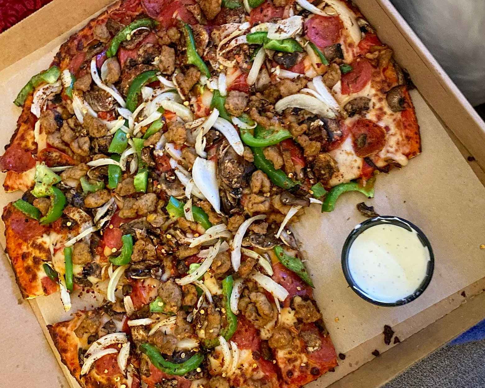 A supreme pizza with a piece taken out, sitting in a pizza box next to a dipping cup of ranch.