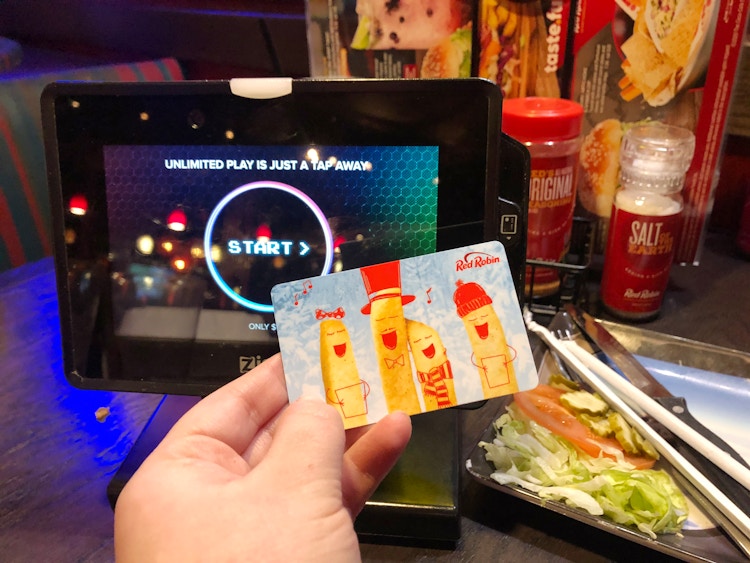 A person's hand holding up a Red Rock gift card in front of a Ziosk machine on a table at Red Robin.