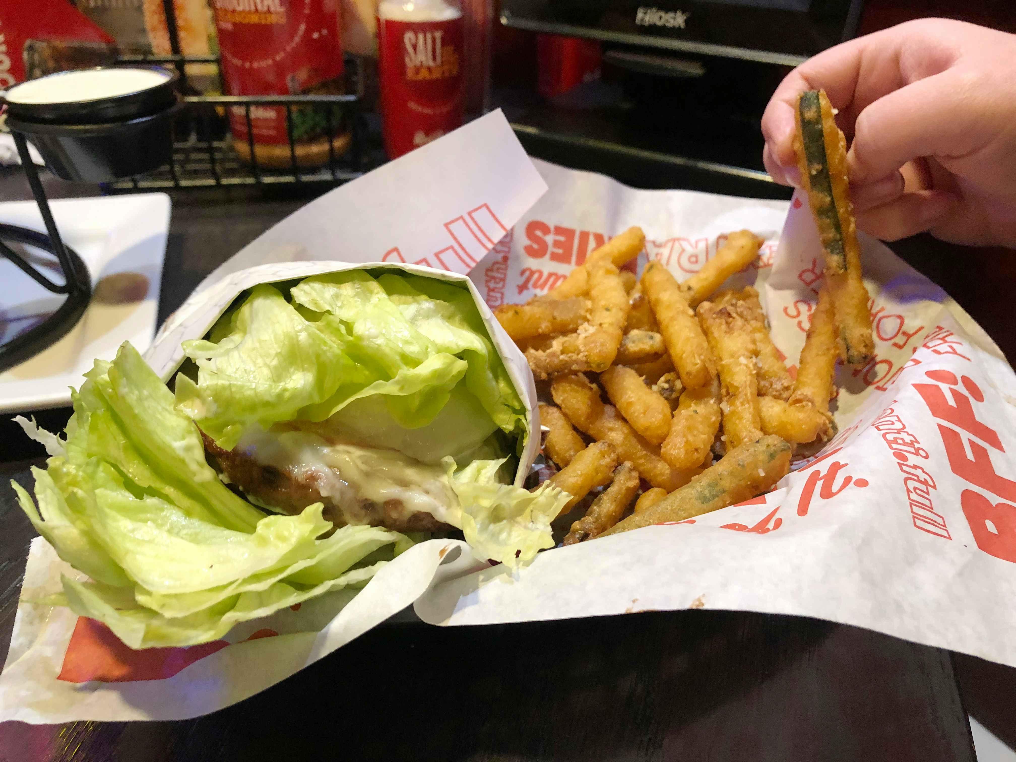 A person's hand taking a zucchini fry from a basket of a lettuce wrapped burger and zucchini fries at Red Robin.