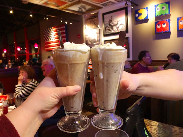 Two people's hands lifting up their milkshakes to cheers inside a Red Robin.