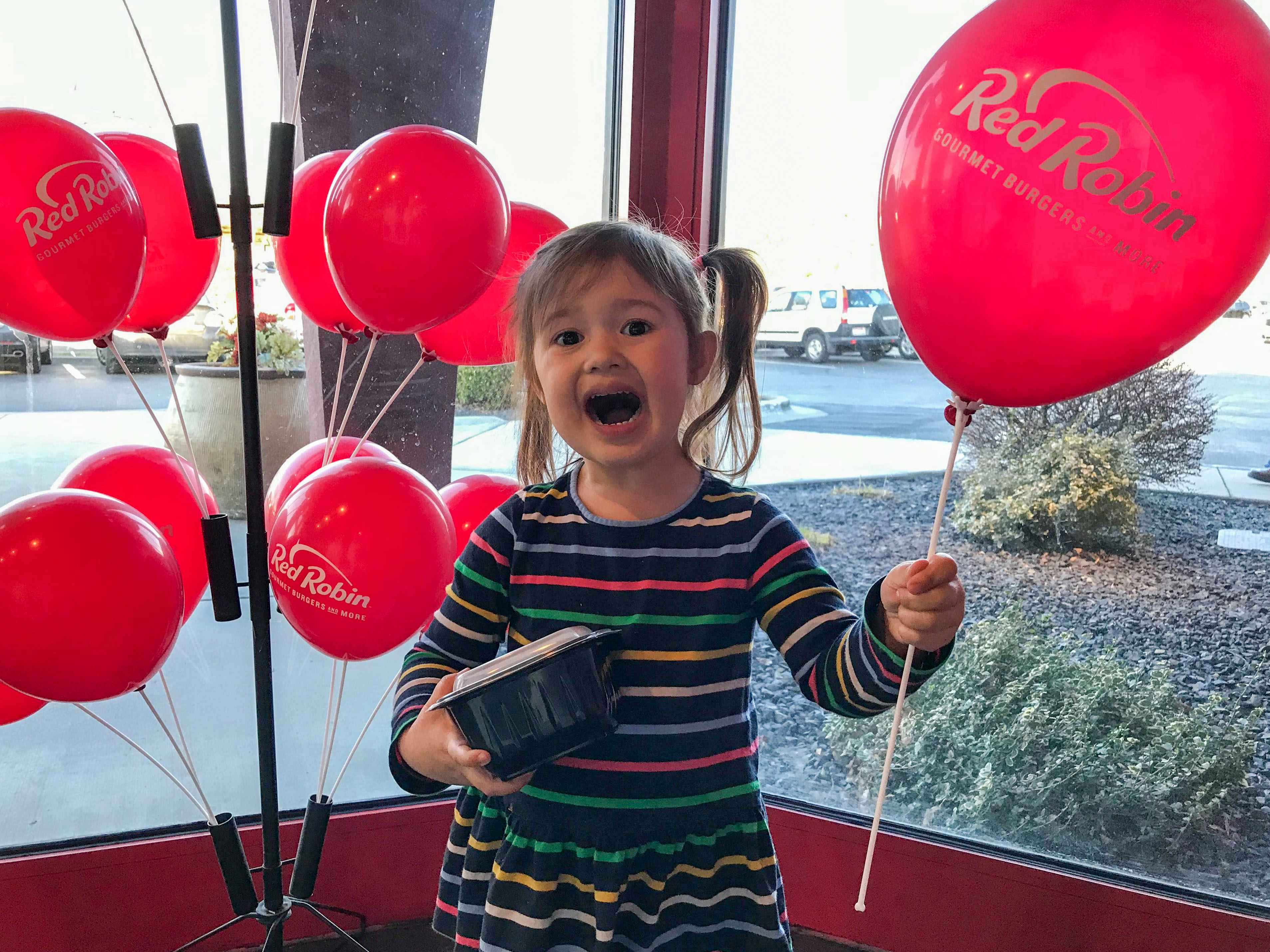 A child inside Red Robin, holding a Red Robin balloon and a takeout container while smiling at the camera.