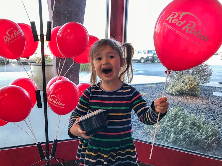 A child inside Red Robin, holding a Red Robin balloon and a takeout container while smiling at the camera.