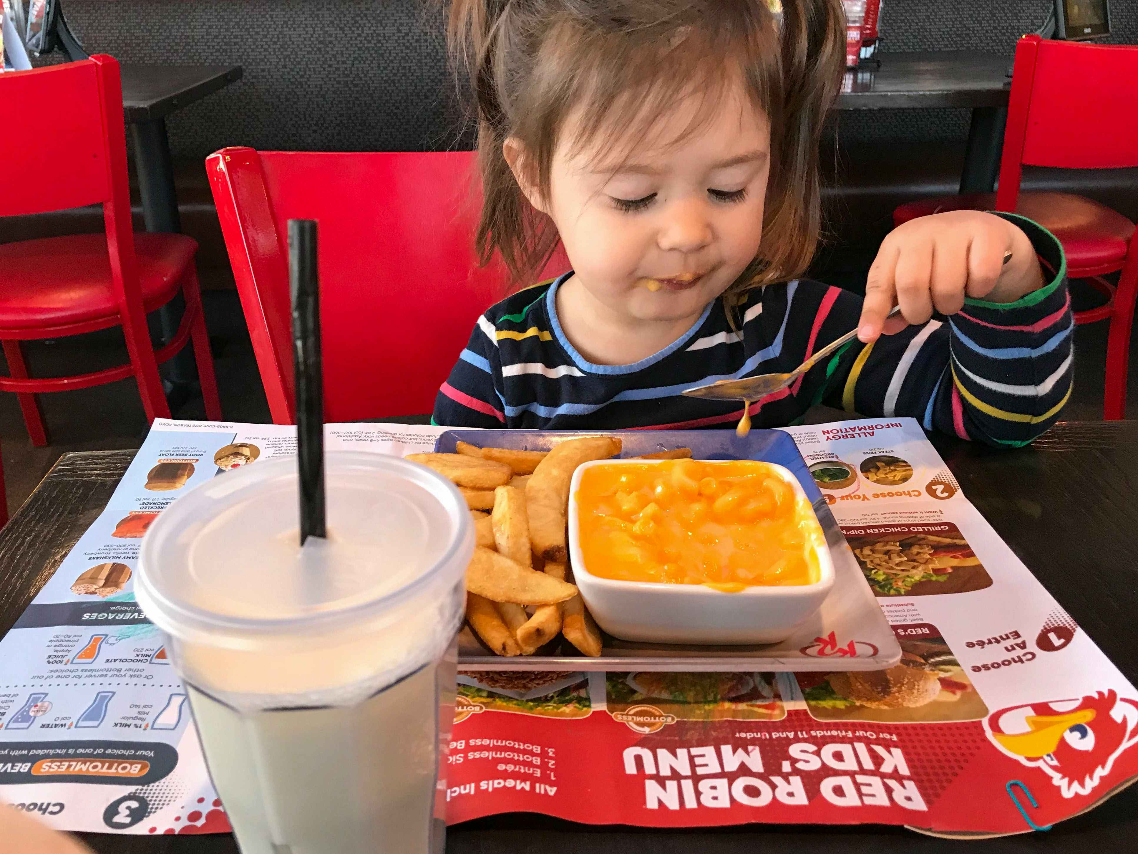 A child sitting at a table at Red Robin, using a spoon to eat mac and cheese. On the table there is a kids' menu spread out and a milkshake.