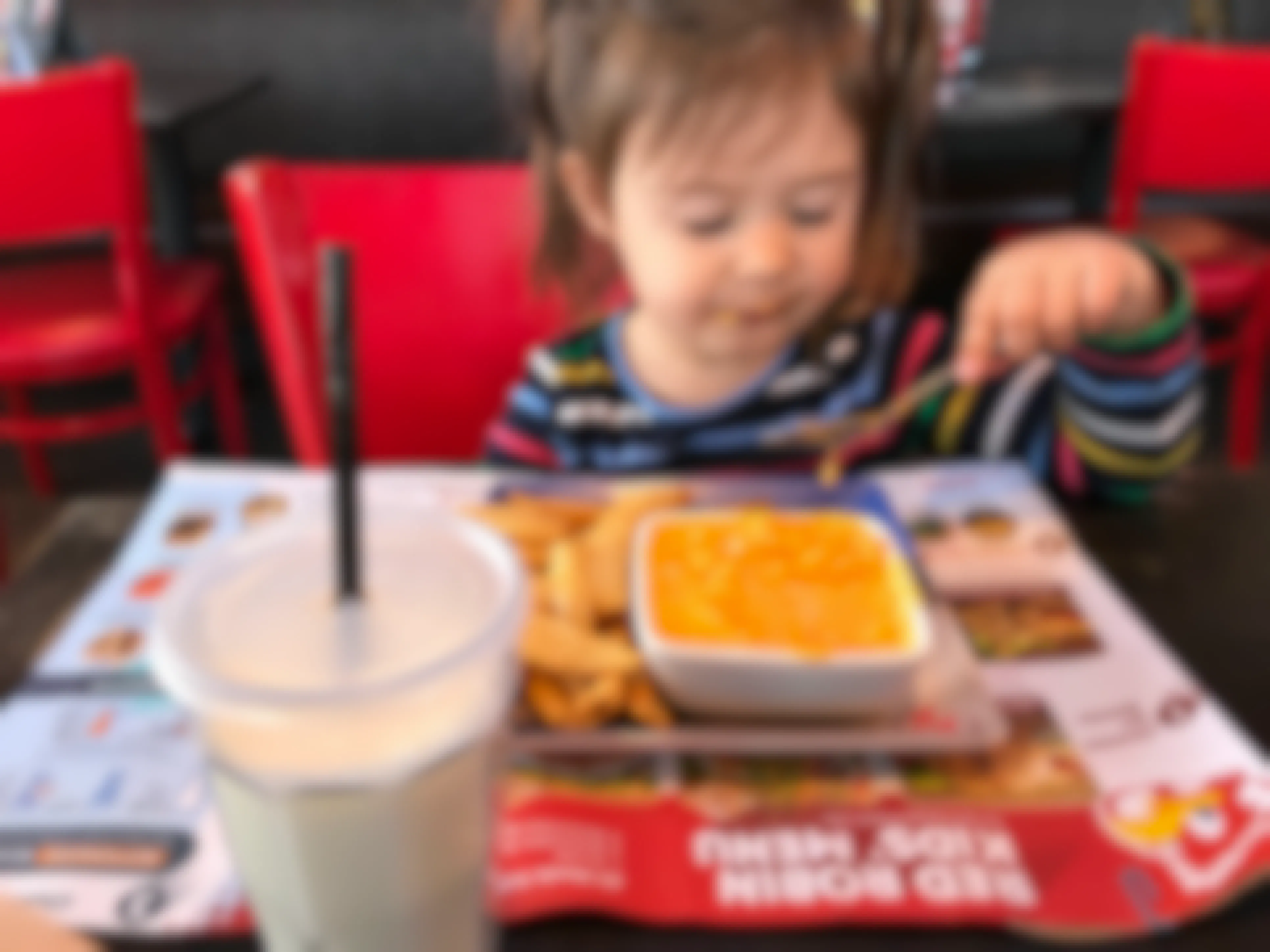 A child sitting at a table at Red Robin, using a spoon to eat mac and cheese. On the table there is a kids' menu spread out and a milkshake.