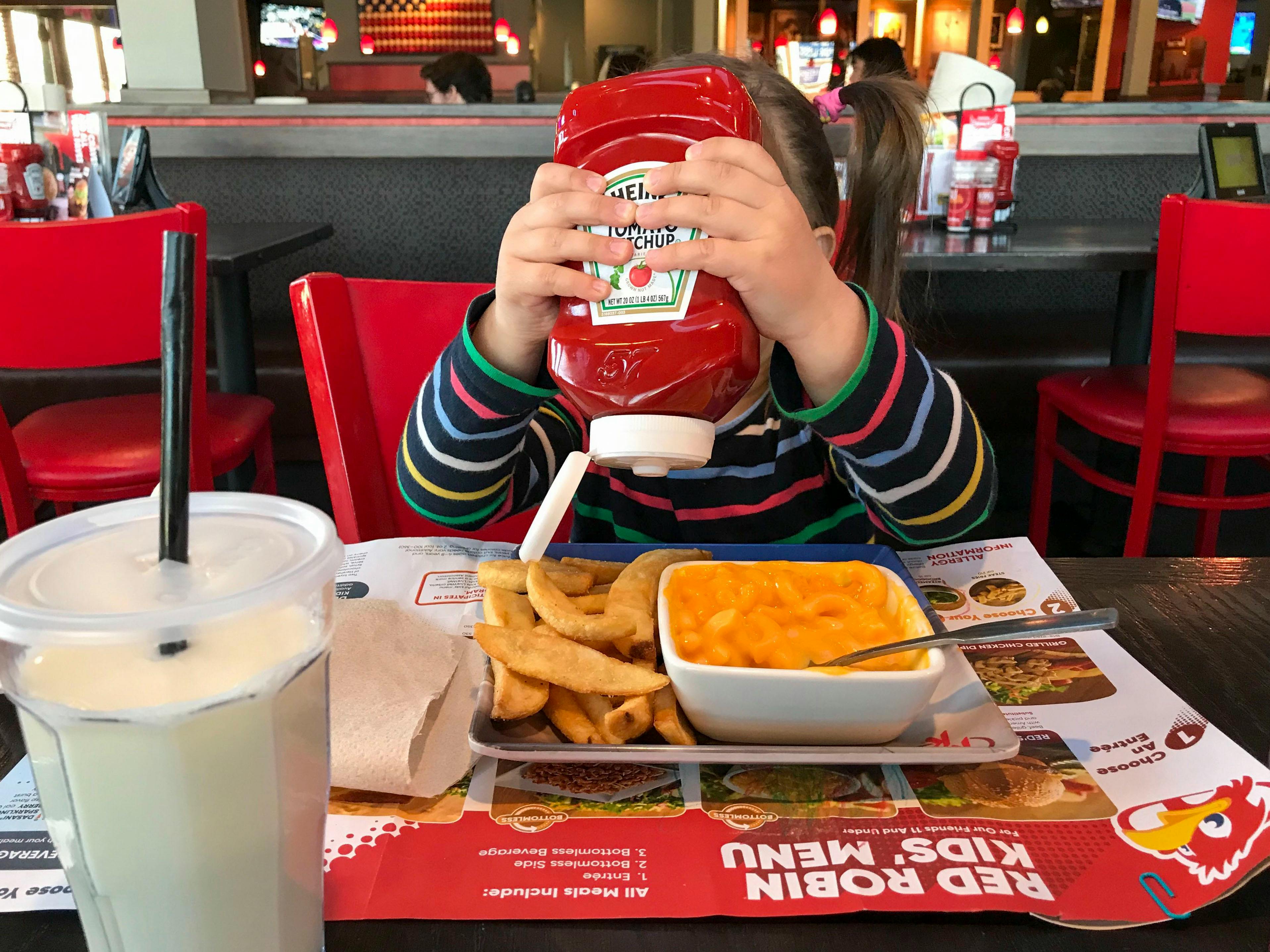 A child squeezing a bottle of Heinz ketchup onto a plate of fries and mac and cheese in front of her at a booth at Red Robin.