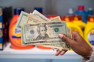 A person holding a stack of cash in front of a stockpile or brand name products in a storage room