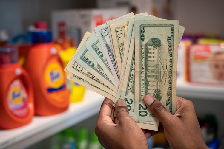 A person holding a stack of cash in front of a stockpile or brand name products in a storage room