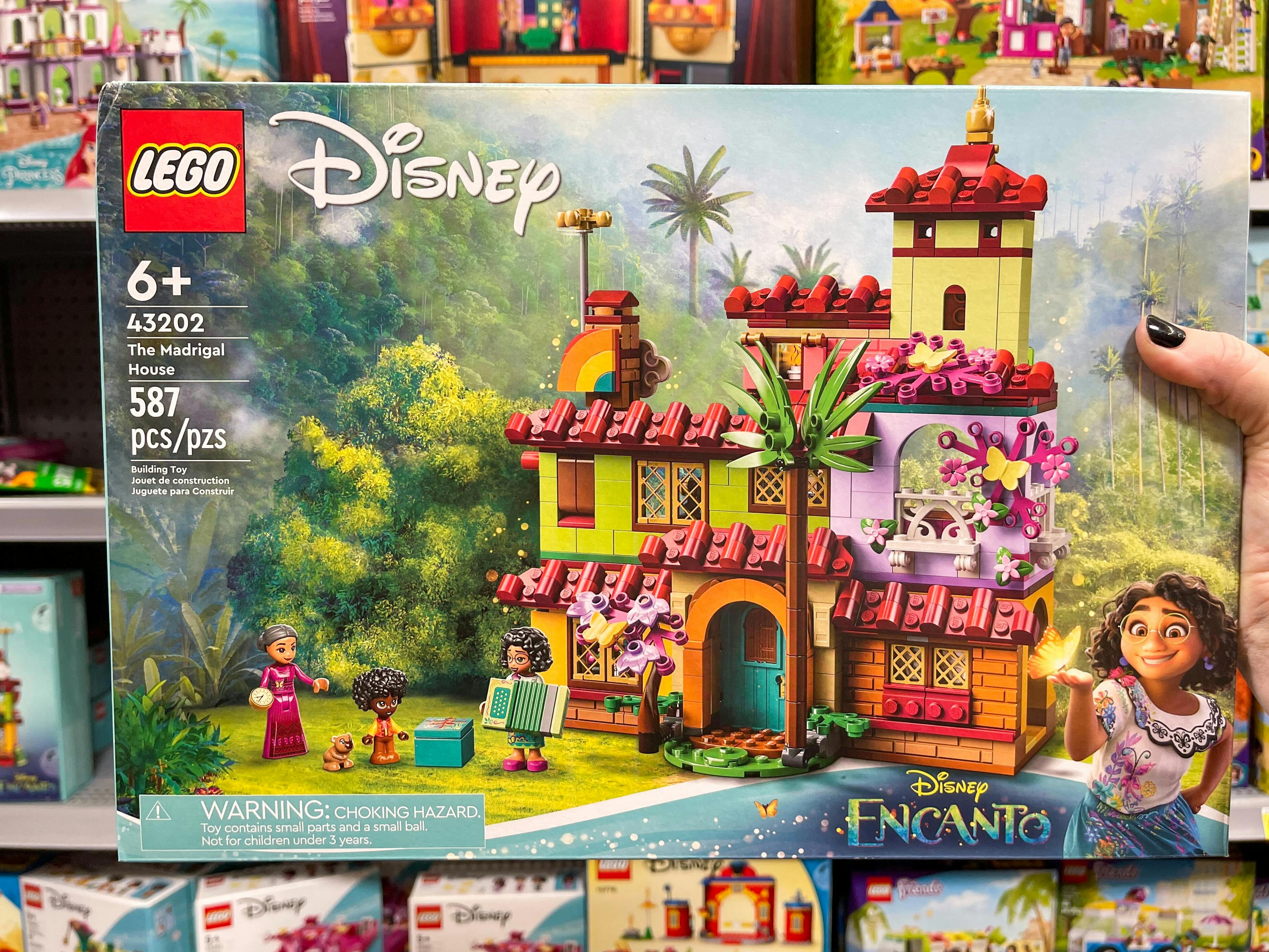 A person's hand holding a LEGO Disney's Encanto Madrigal house building set in front of a shelf of toys.