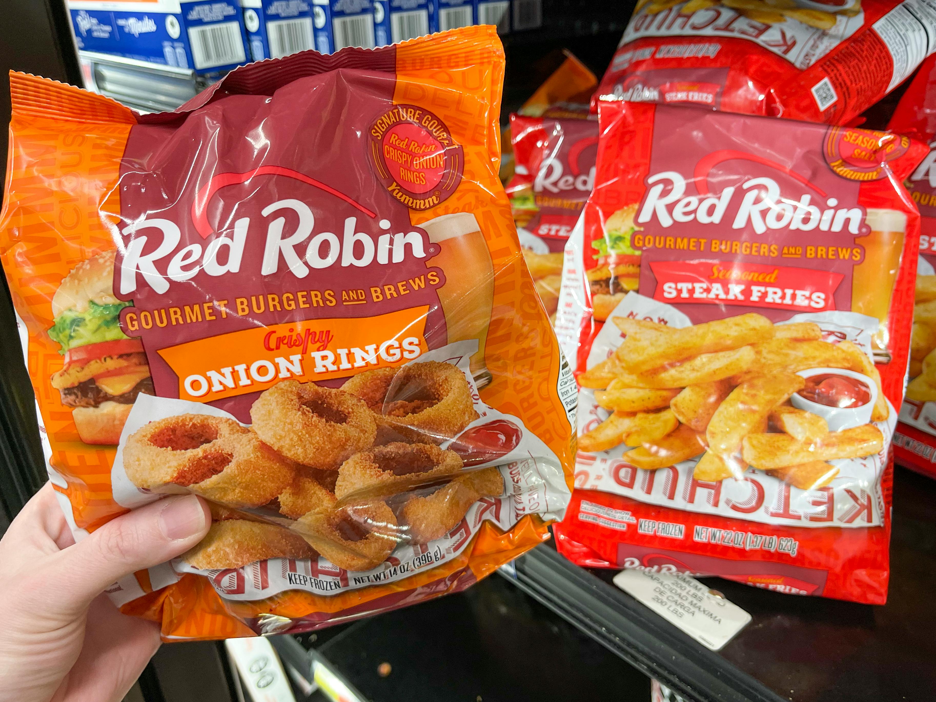 A person's hand holding up Red Robin onion rings next to a bag of Red Robin french fries in the freezer section at Walmart.