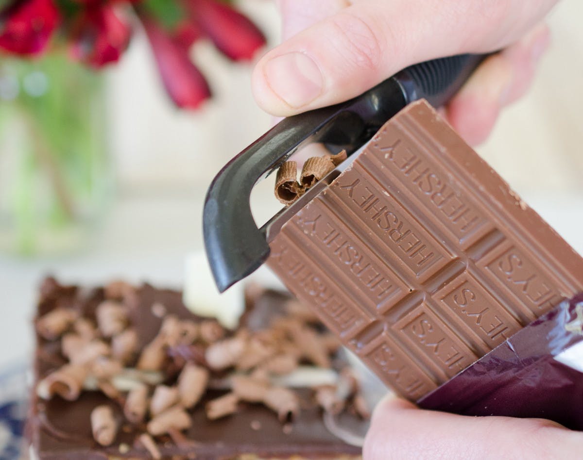 A person using a potato peeler to peel a chocolate bar into chocolate shavings.