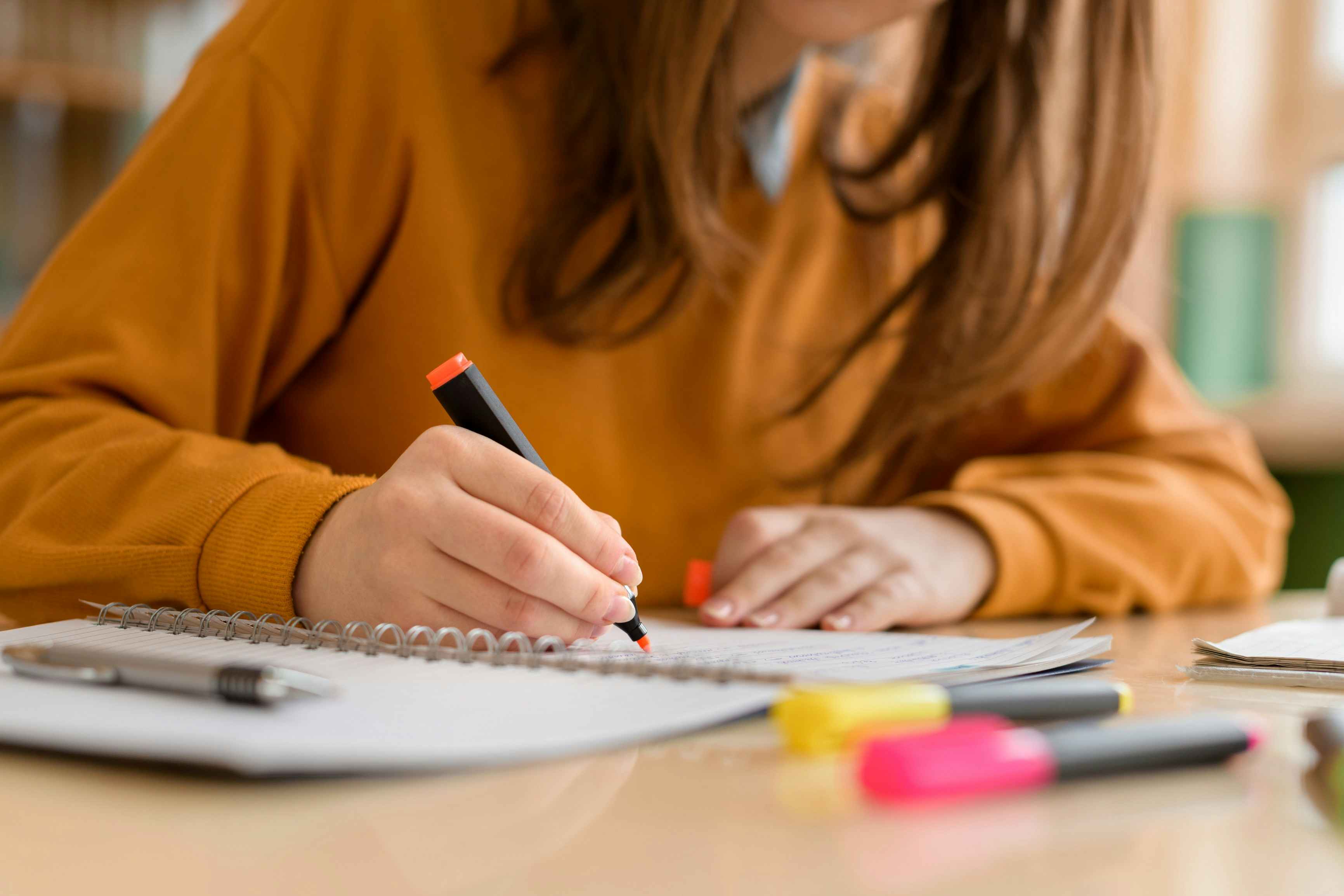 Girl in an orange sweater with a highlighter in her hand staring at a notebook