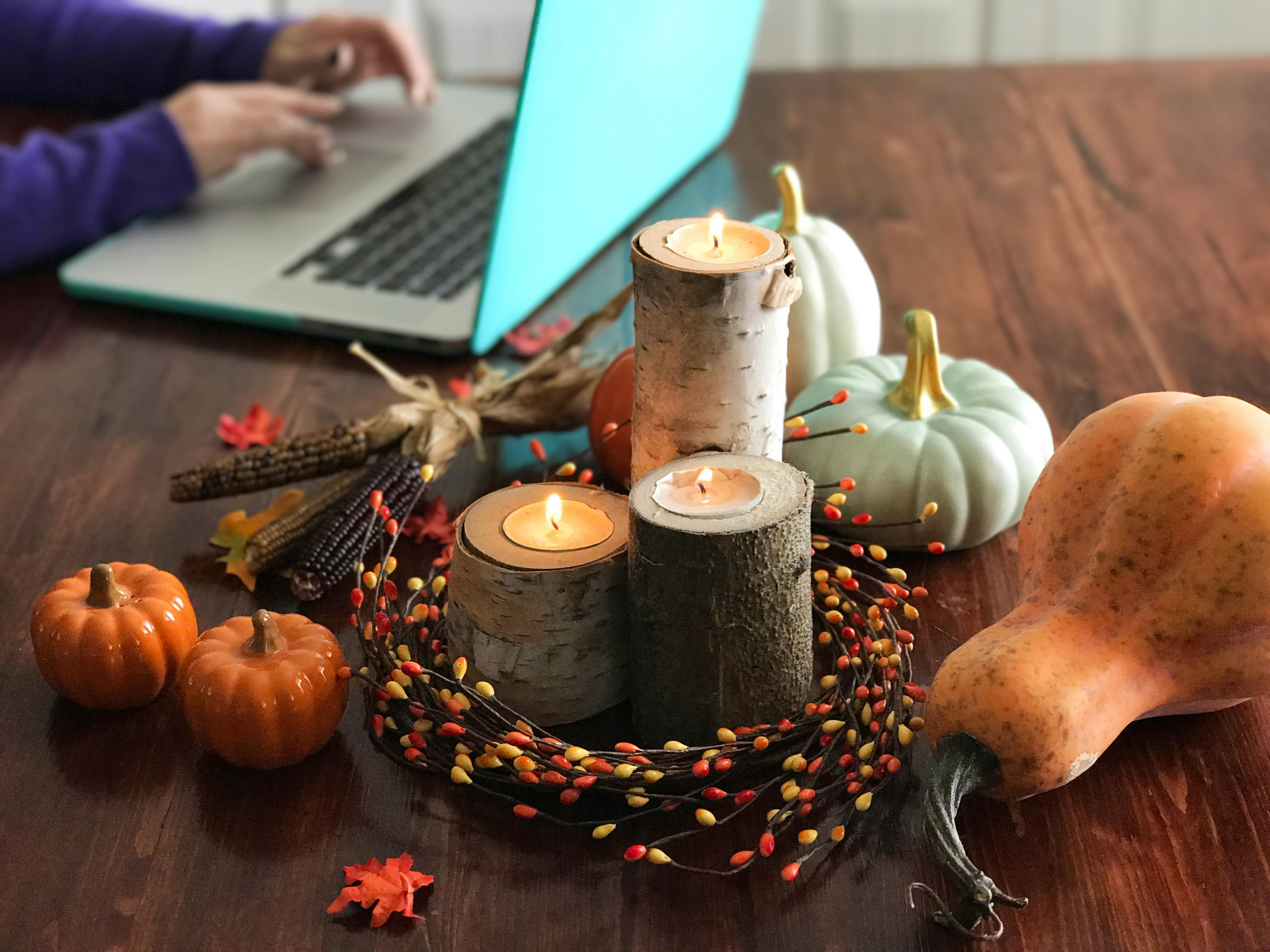 A autumn candle centerpiece on a table with gourds next to someone using a laptop.