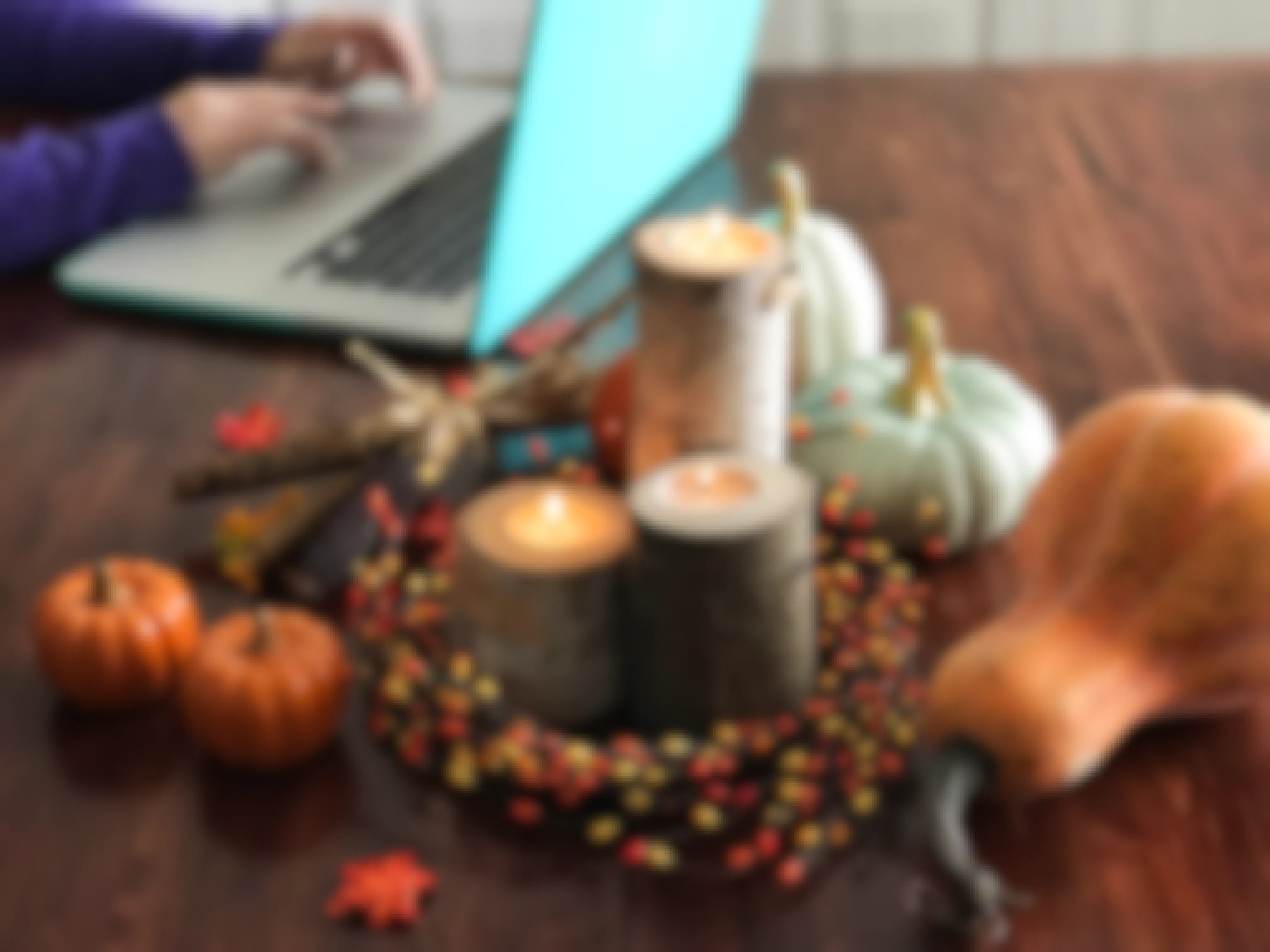 A autumn candle centerpiece on a table with gourds next to someone using a laptop.