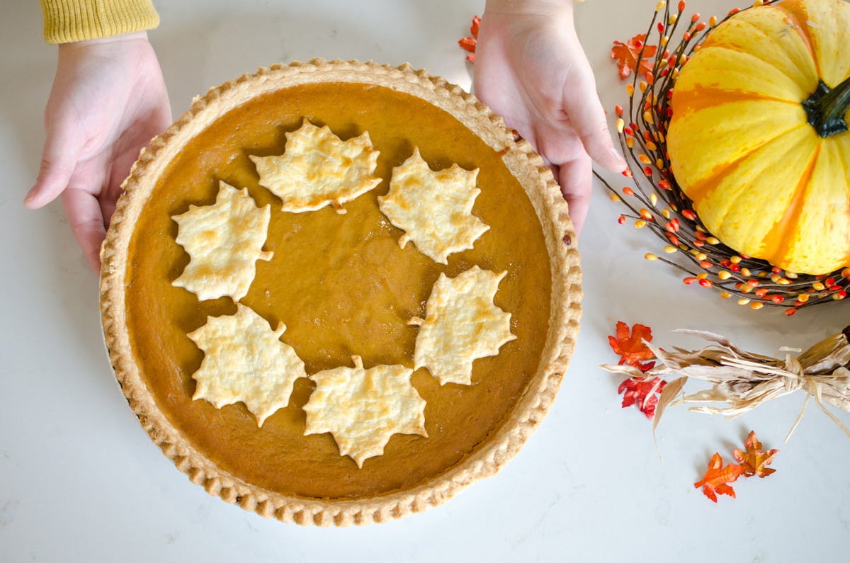 A pumpkin pie decorated with autumn leaves made of pie crust being set down on a table next to a pumpkin decoration.
