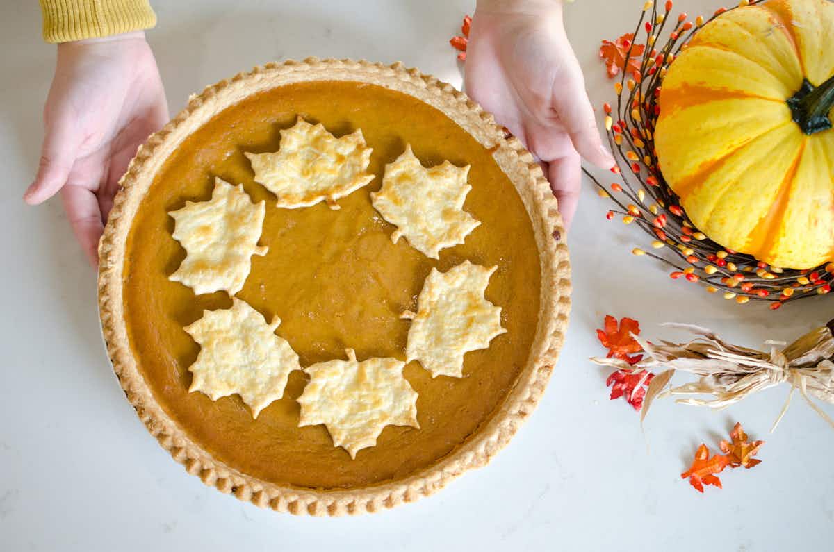 A pumpkin pie decorated with autumn leaves made of pie crust being set down on a table next to a pumpkin decoration.