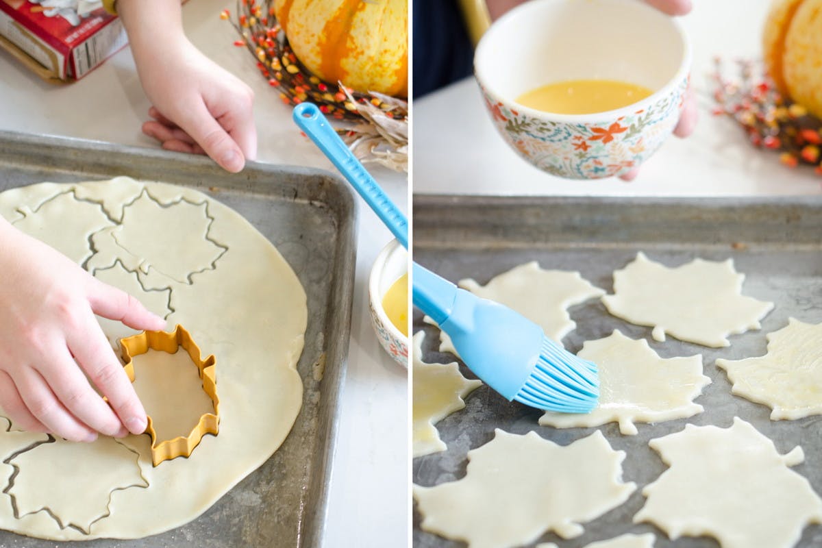 A person using a leaf-shaped cookie cutter to cut leaf shapes out of pie crust dough and using a basting brush to cover them with egg wash.