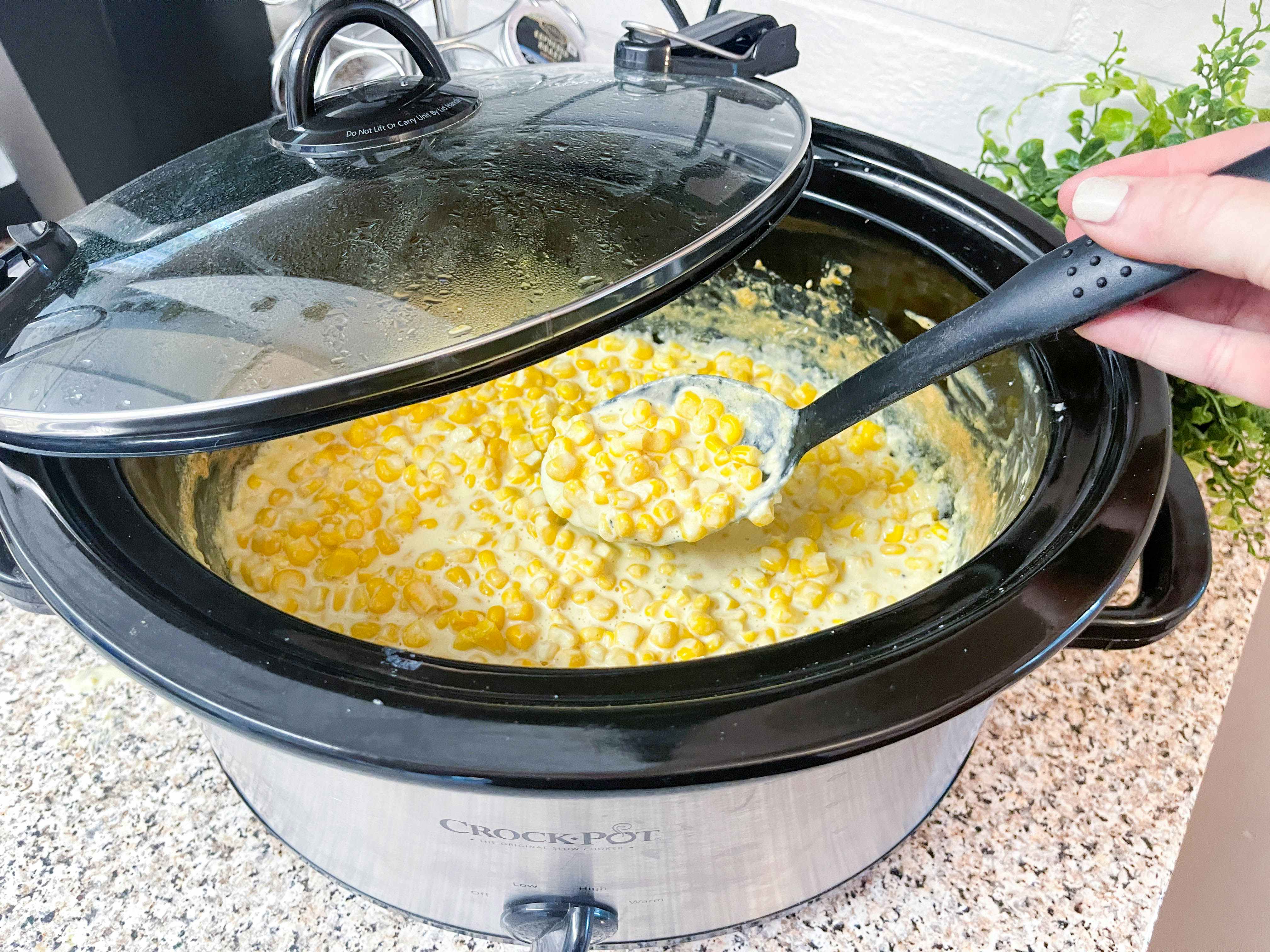 A slow cooker filled with cooked creamed corn with a person scooping it out..