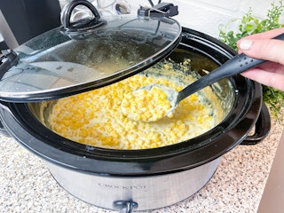 A slow cooker filled with cooked creamed corn with a person scooping it out..