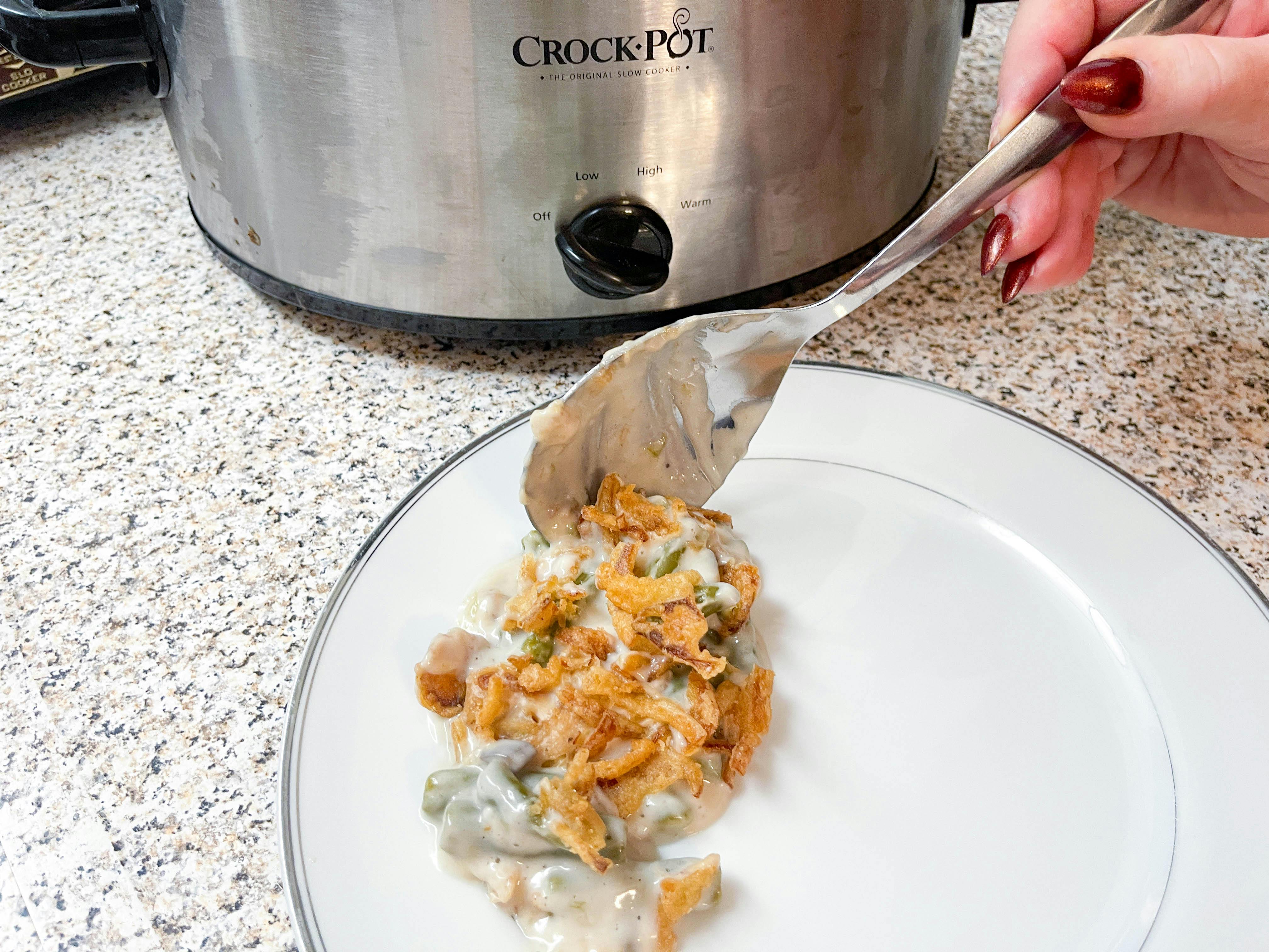 A spoon full of green beans casserole being put on a plate in front of a crock pot.