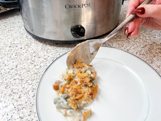 A spoon full of green beans casserole being put on a plate in front of a crock pot.