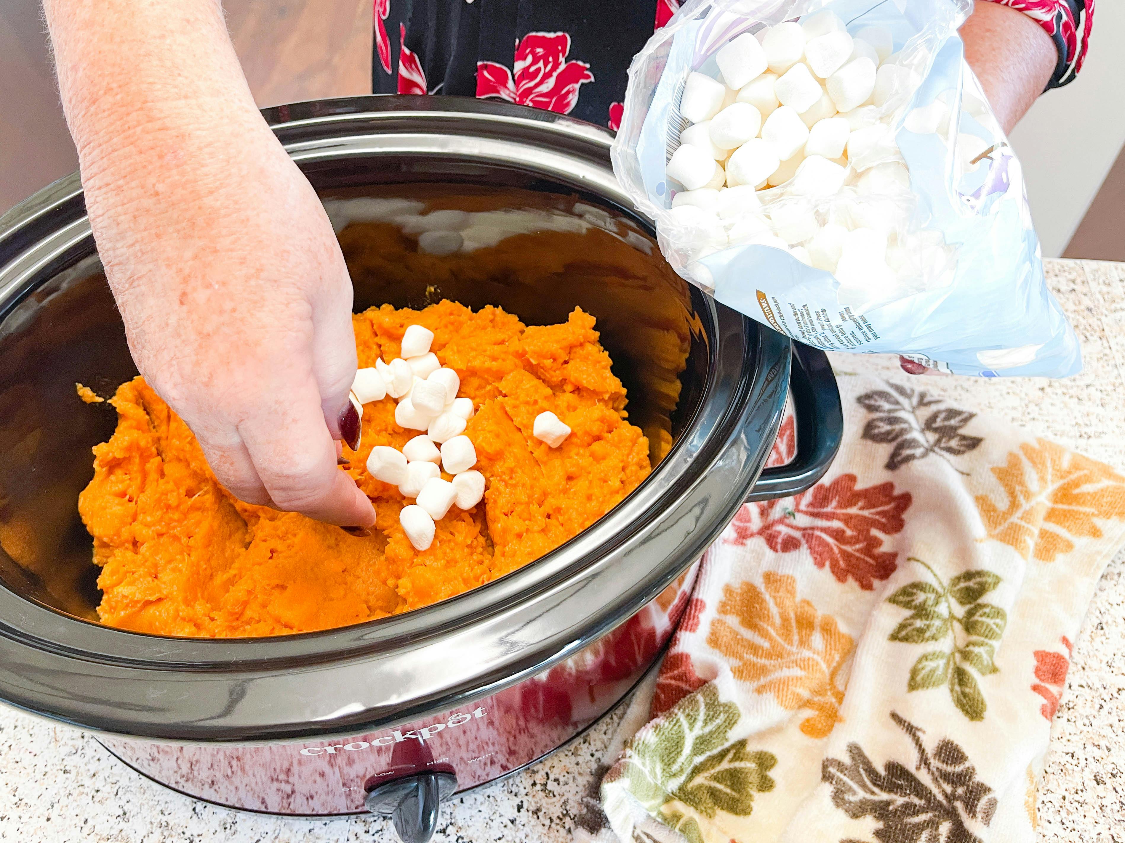 A crockpot filled with sweet potatoes with a person placing mini marshmallows on top