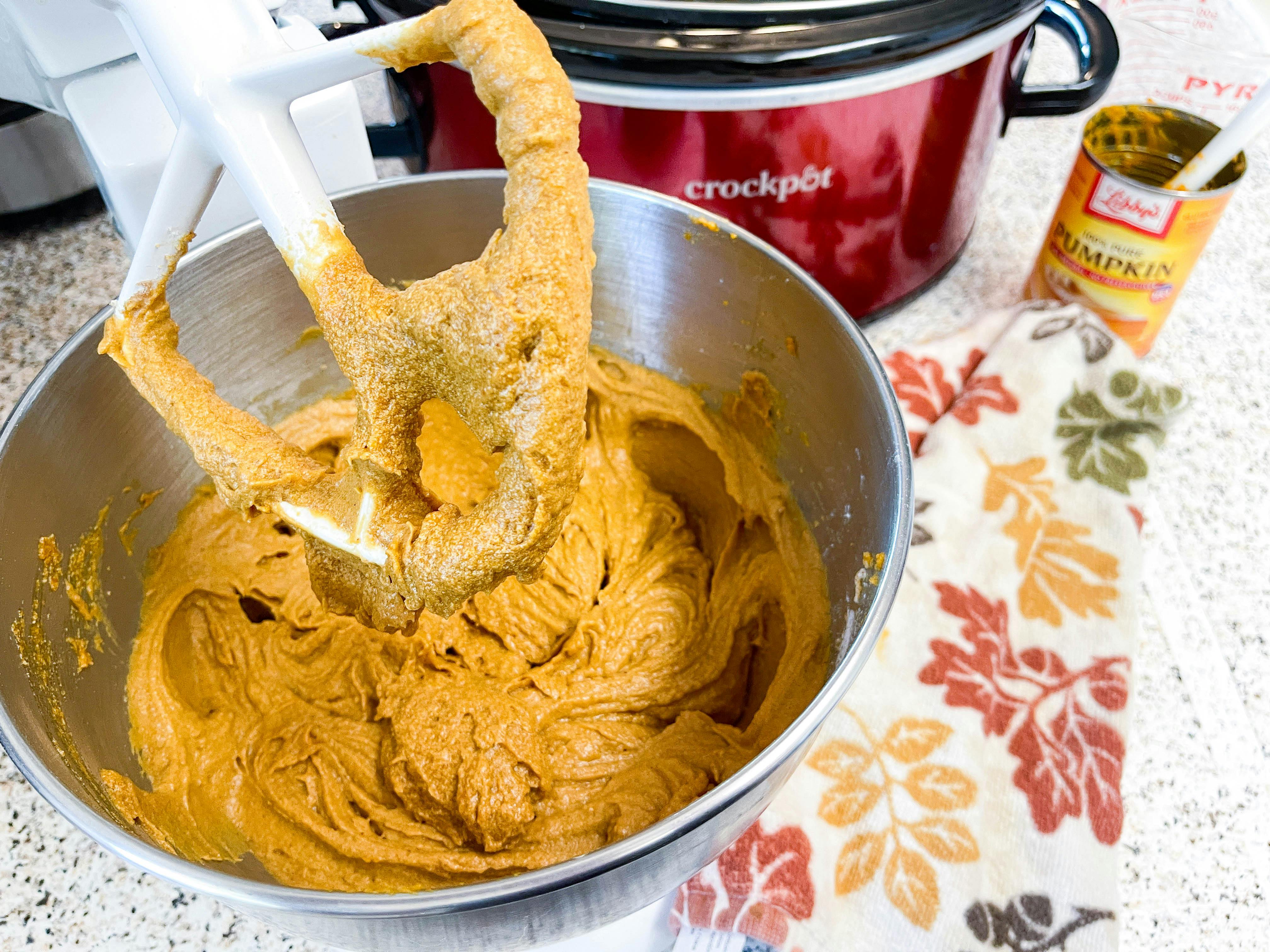 A mixer beater with pumpkin cake batter in the bowl next to a slow cooker.