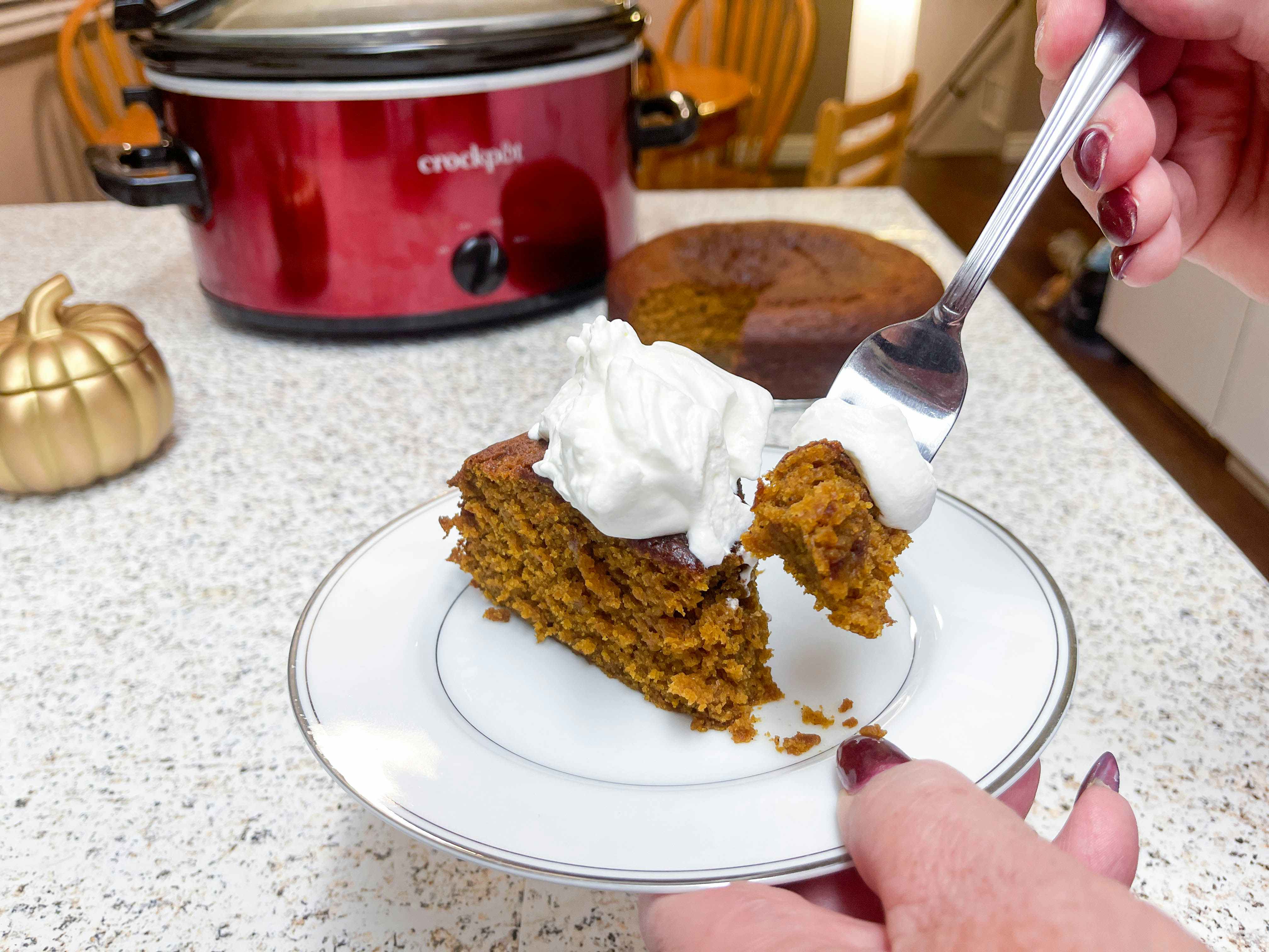 A slice of pumpkin cake on a plate with a person scooping a piece on a fork in front of a crock pot