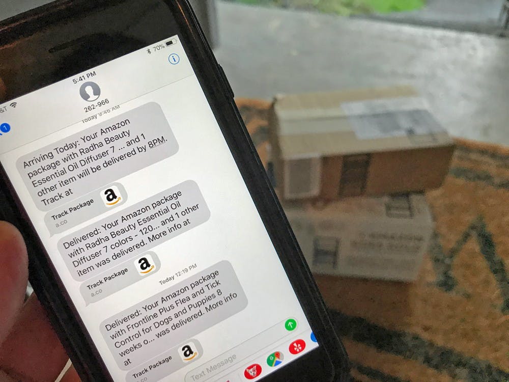 A person's hand holding their phone displaying the Amazon shipment updates they received via text, with two packages stacked on their porch in the background.