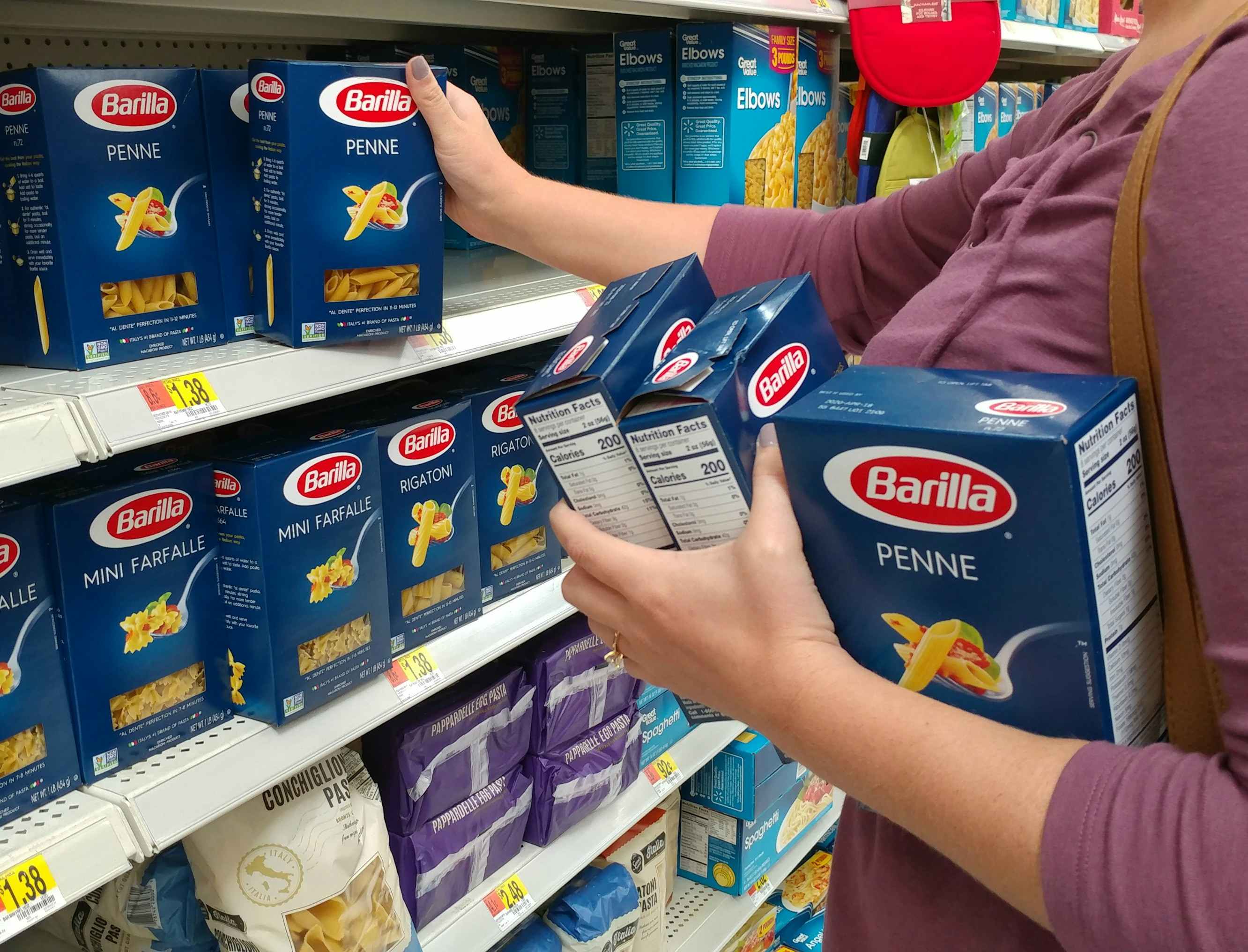 A woman taking Barilla Penne pasta off a walmart grocery store shelf with 3 boxes already in her other arm.