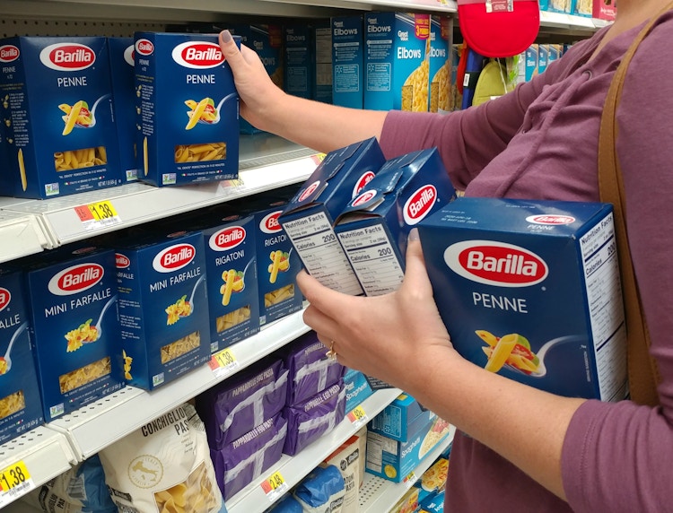 A woman taking Barilla Penne pasta off a walmart grocery store shelf with 3 boxes already in her other arm.