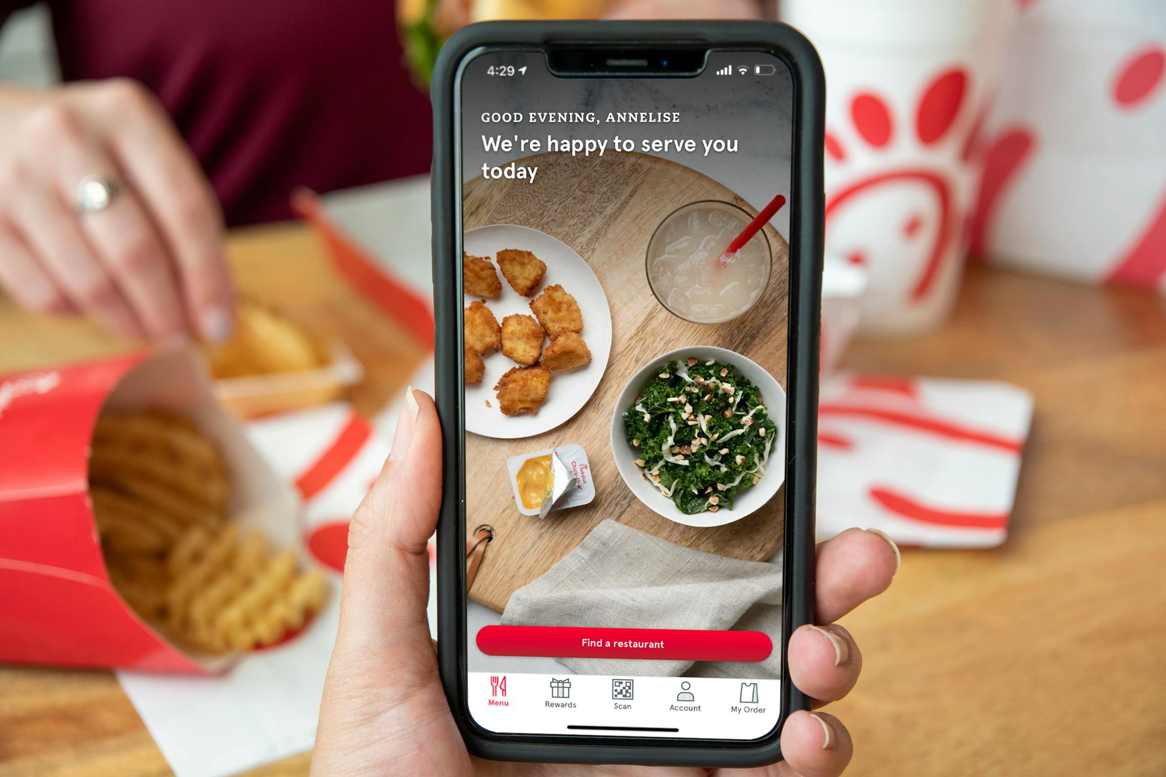 The Chick-fil-a app on a cell phone with a person eating chick-fil-a in the background.