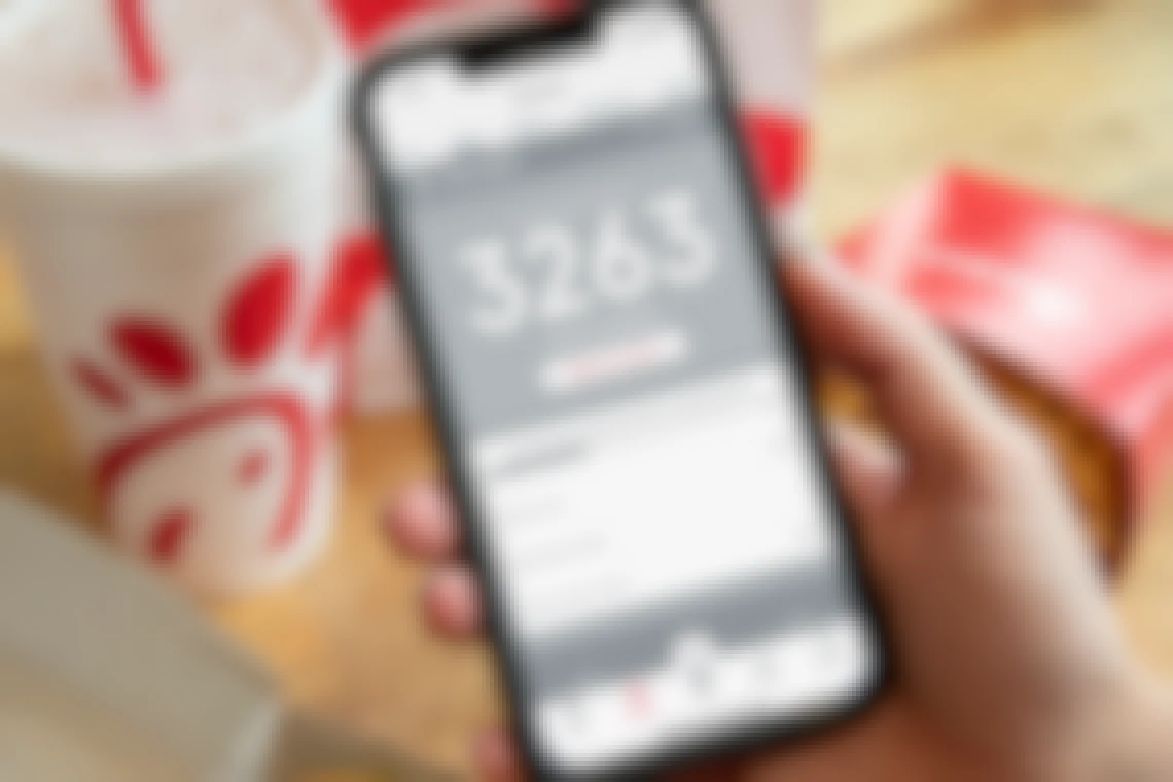A person's hand holding an iPhone displaying the Points tab of the Chick-fil-A mobile app with a box of Chick-fil-A waffle fries, a takeout bag, and a fountain drink cup on the table in the background.