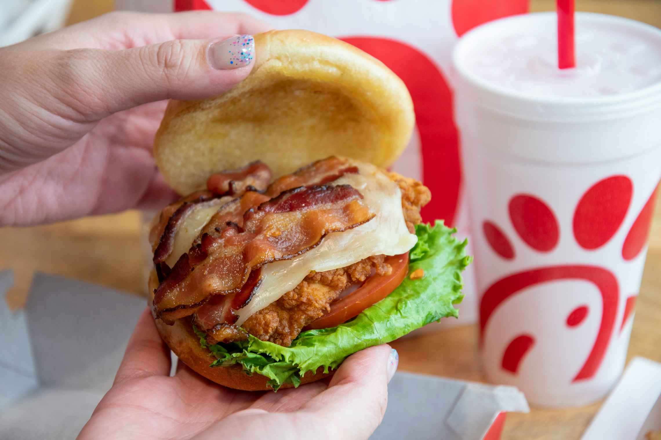 A person's hands lifting the top bun off of a Chick-fil-A chicken sandwich with bacon to show the ingredients. On the table in the background is a Chick-fil-A bag and drink cup.