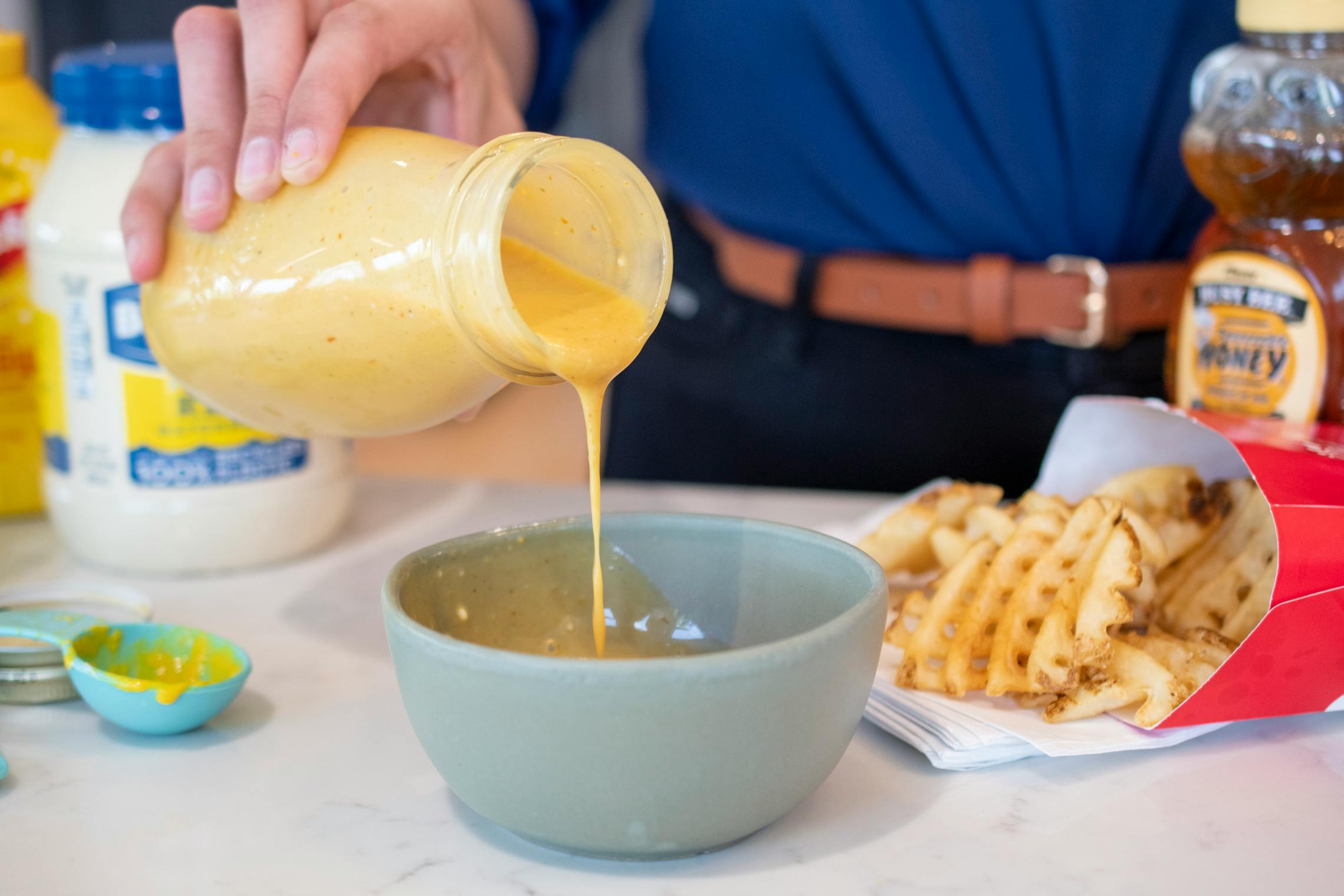 Homemade chick-fil-a sauce being poured from a glass bottle into a bowl on a table next to Chick-fil-A waffle fries and sauce ingredients.