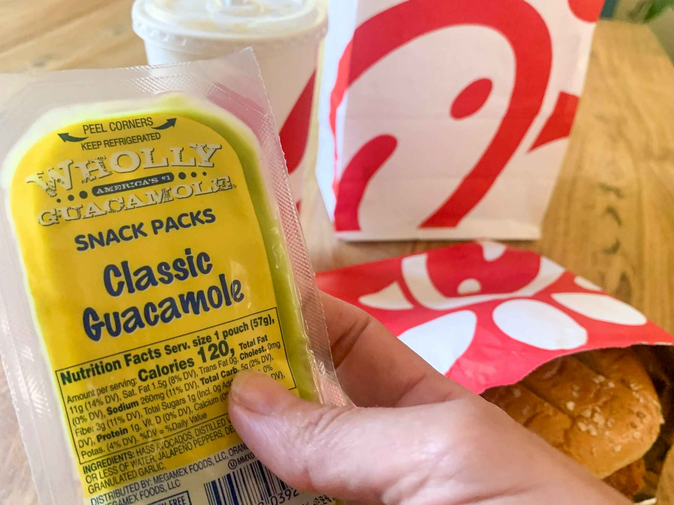 A person's hand holding a snack-pack-sized classic Guacamole next to a Chick-fil-A sandwich and drink.