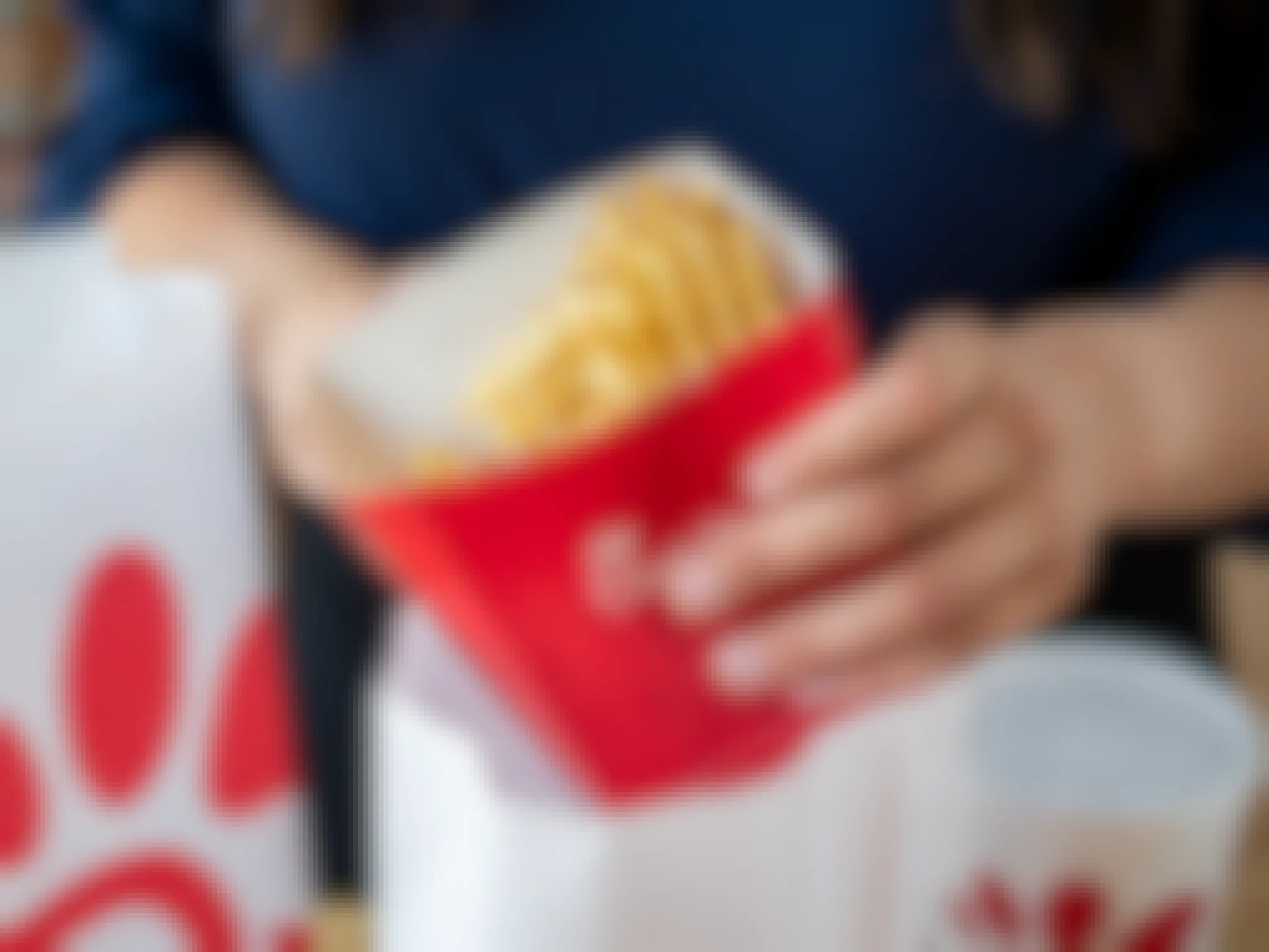A person's hand taking a box of Chick-fil-A waffle fries out of a Chick-fil-A takeout bag sitting on a table next to another Chick-fil-A takeout bag and a fountain drink cup.