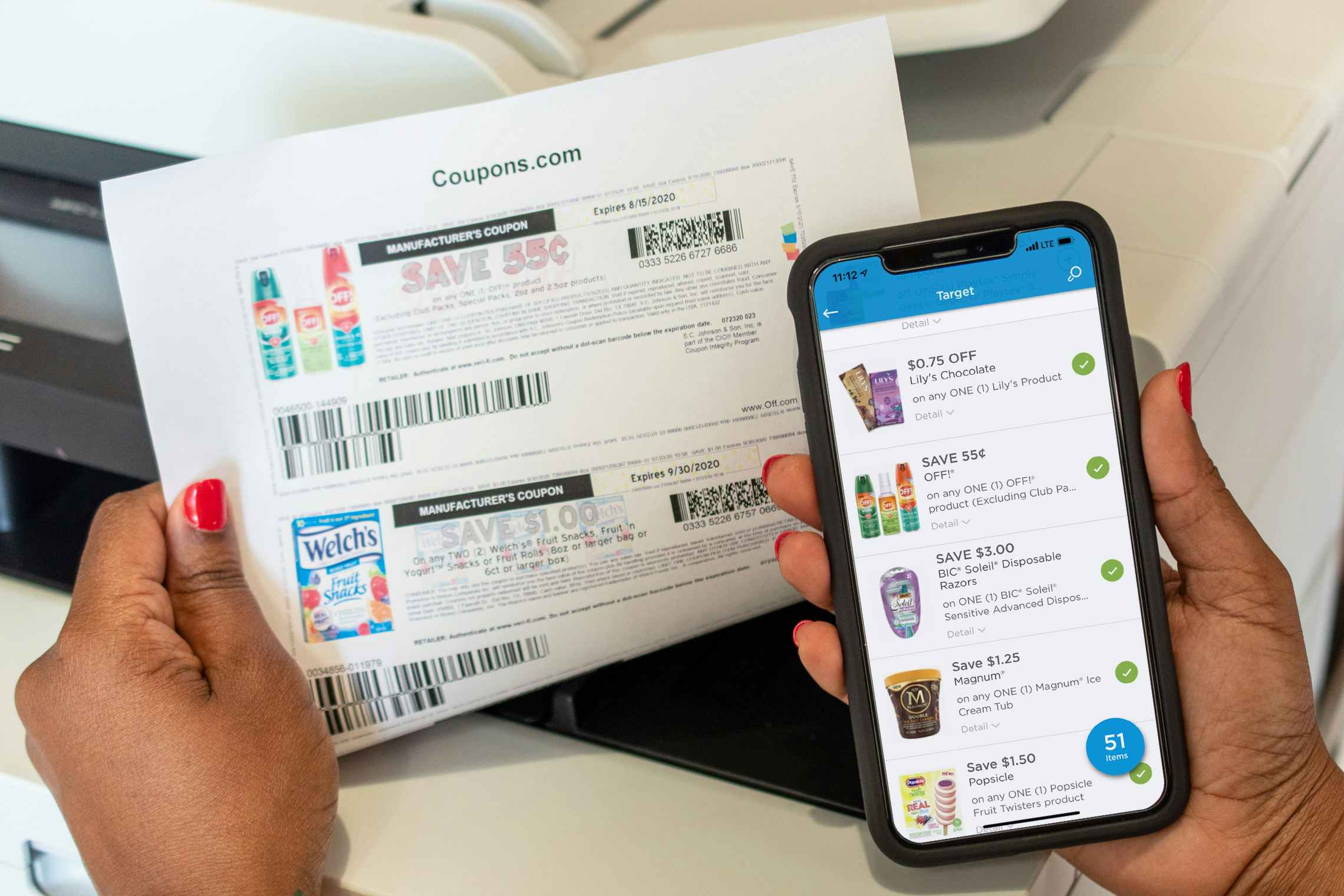 A person holding a cell phone displaying the coupons.com app and printed coupons in front of a printer.