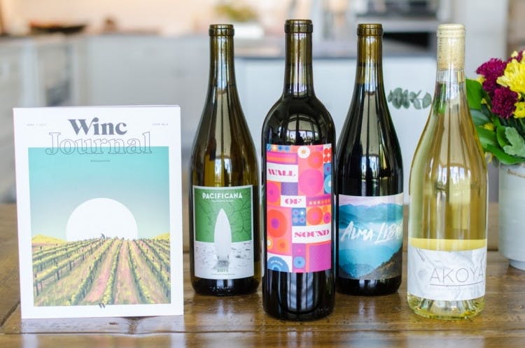 four bottles of wine on a table standing next to a card with the Winc logo on italong with a horizon over a field.