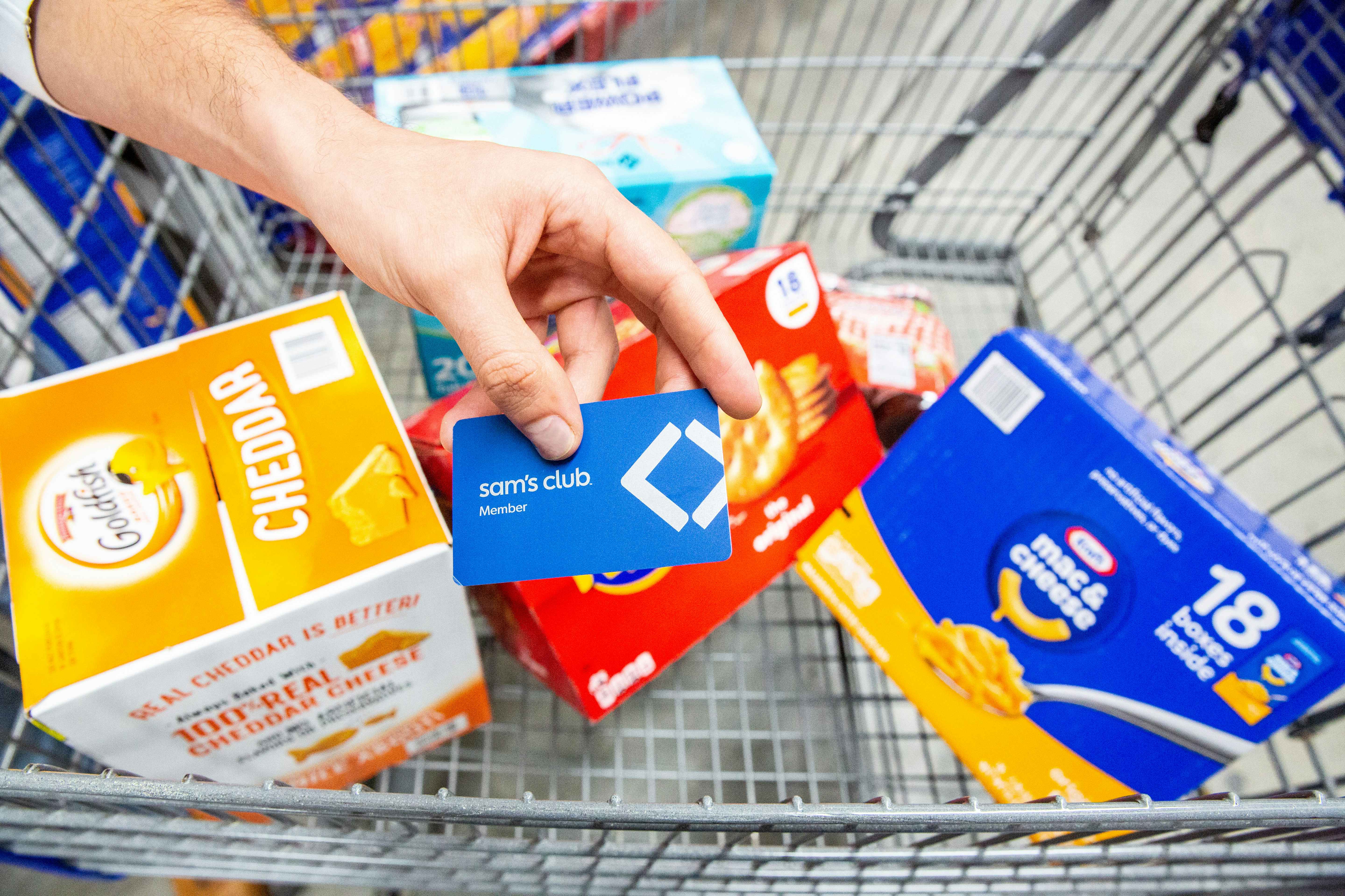 Hand holding a blue Sam's Club membership card above bulk boxes of various snacks like goldfish, kraft mac and cheese, and ritz crackers