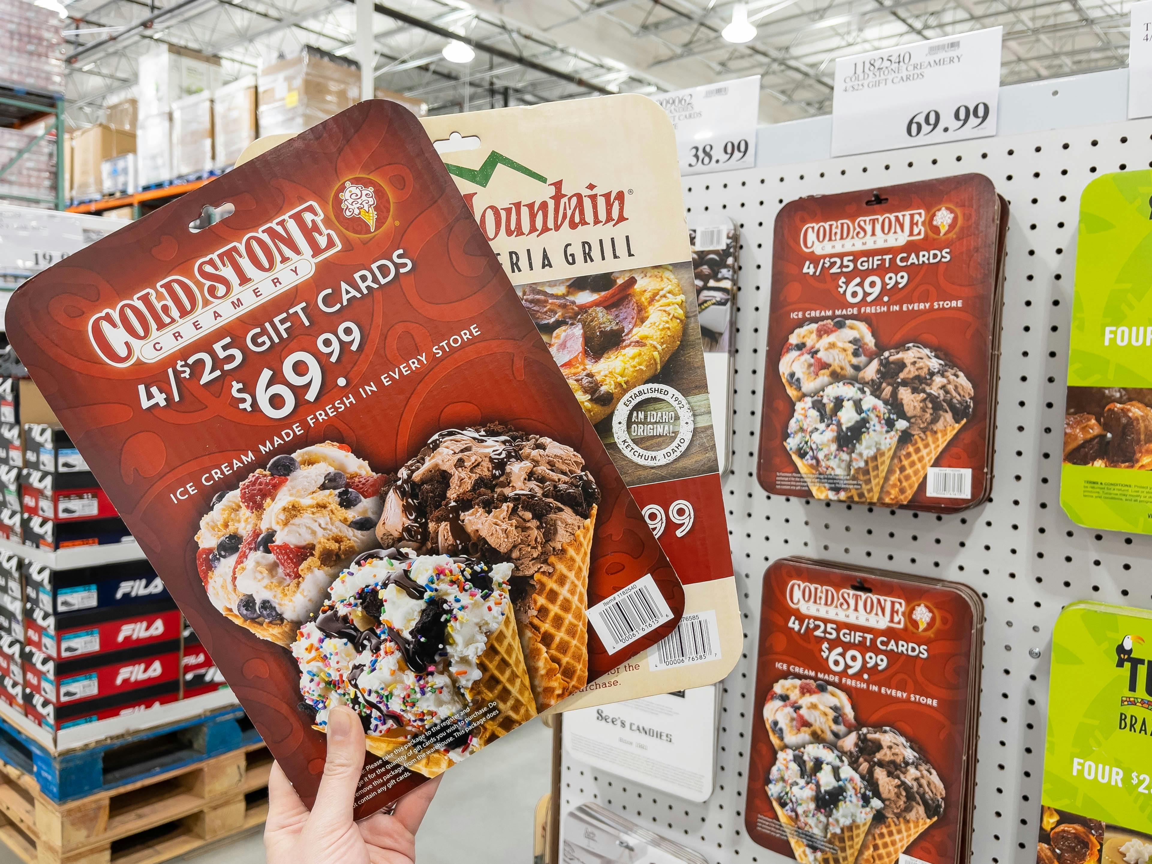 A person holding up two cardboard display cutouts for gift cards sold at Costco. One of the cards is for 4, $25 Cold Stone Creamery gift cards for $69.99