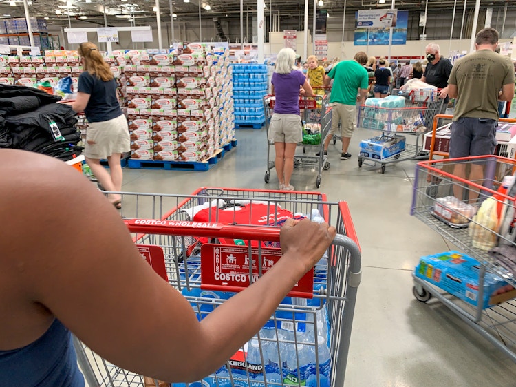 A woman from behind, pushing a Costco shopping cart through a crowd of other shoppers also pushing carts.