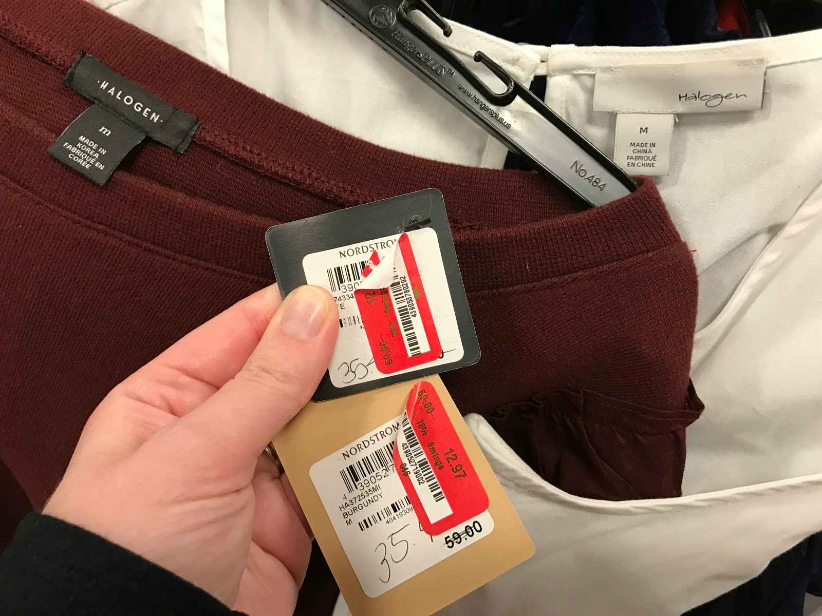 A person holding two price tags attached to clothing. Each tag has a red and white price sticker on it. Those stickers a peeled back to reveal the Nordstrom label on the tag. 