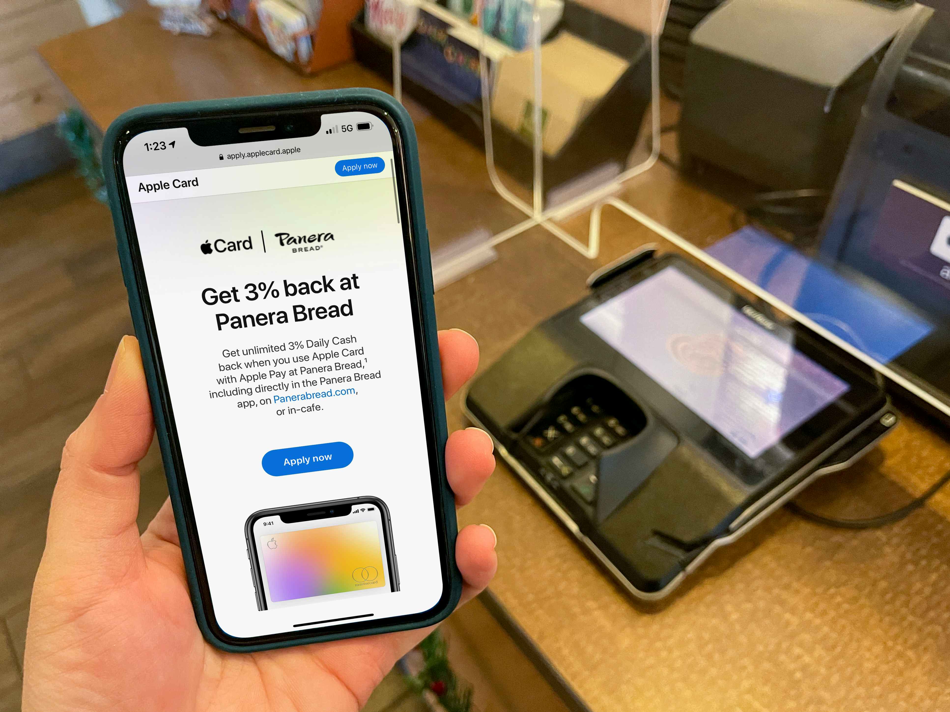 A cell phone held next to a Panera register showing a deal for 3% back at Panera with Apple Pay.